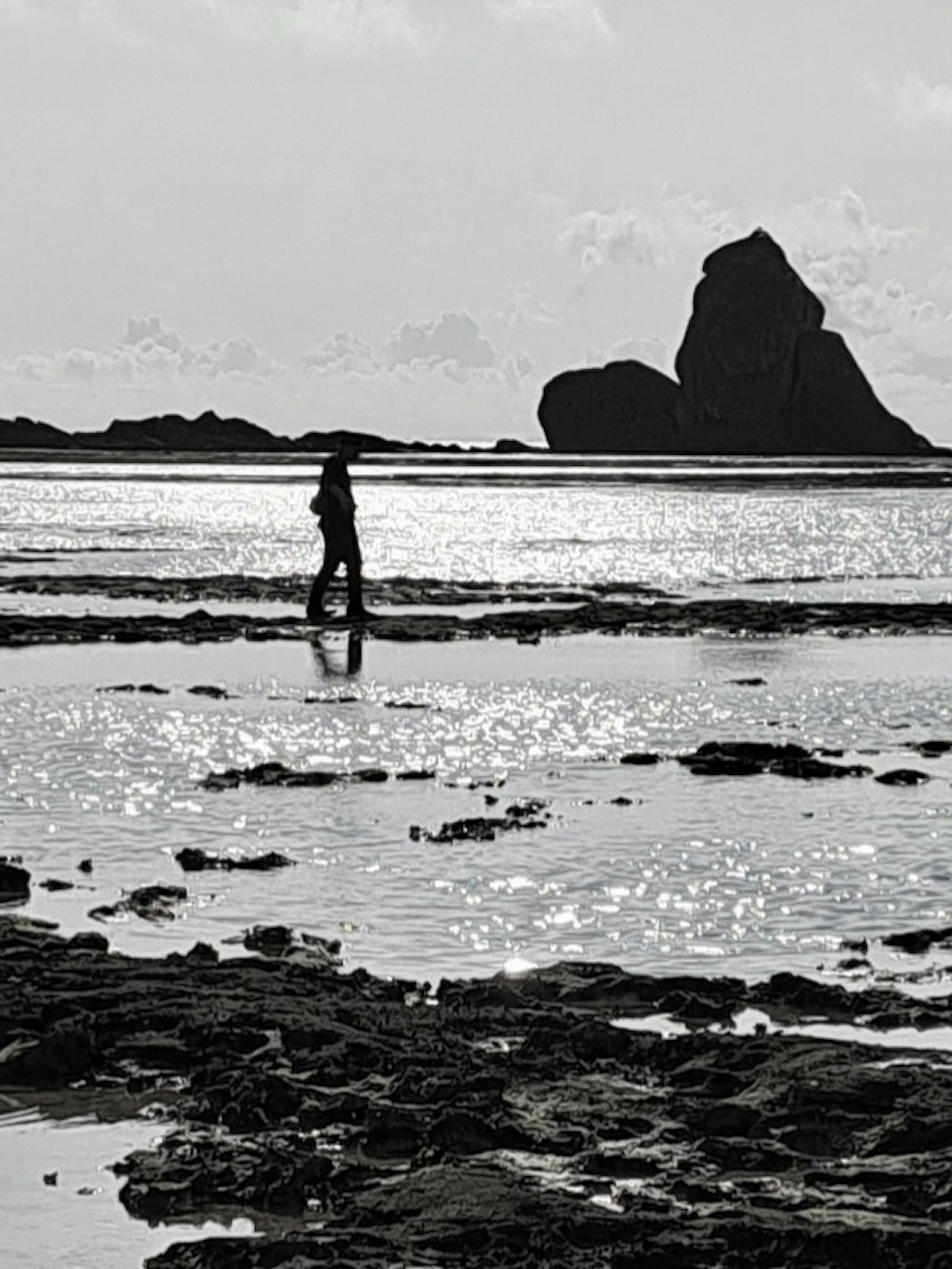 Silhouette of a person walking on a tidal flat with a large rock in the background reflecting sunlight on the water