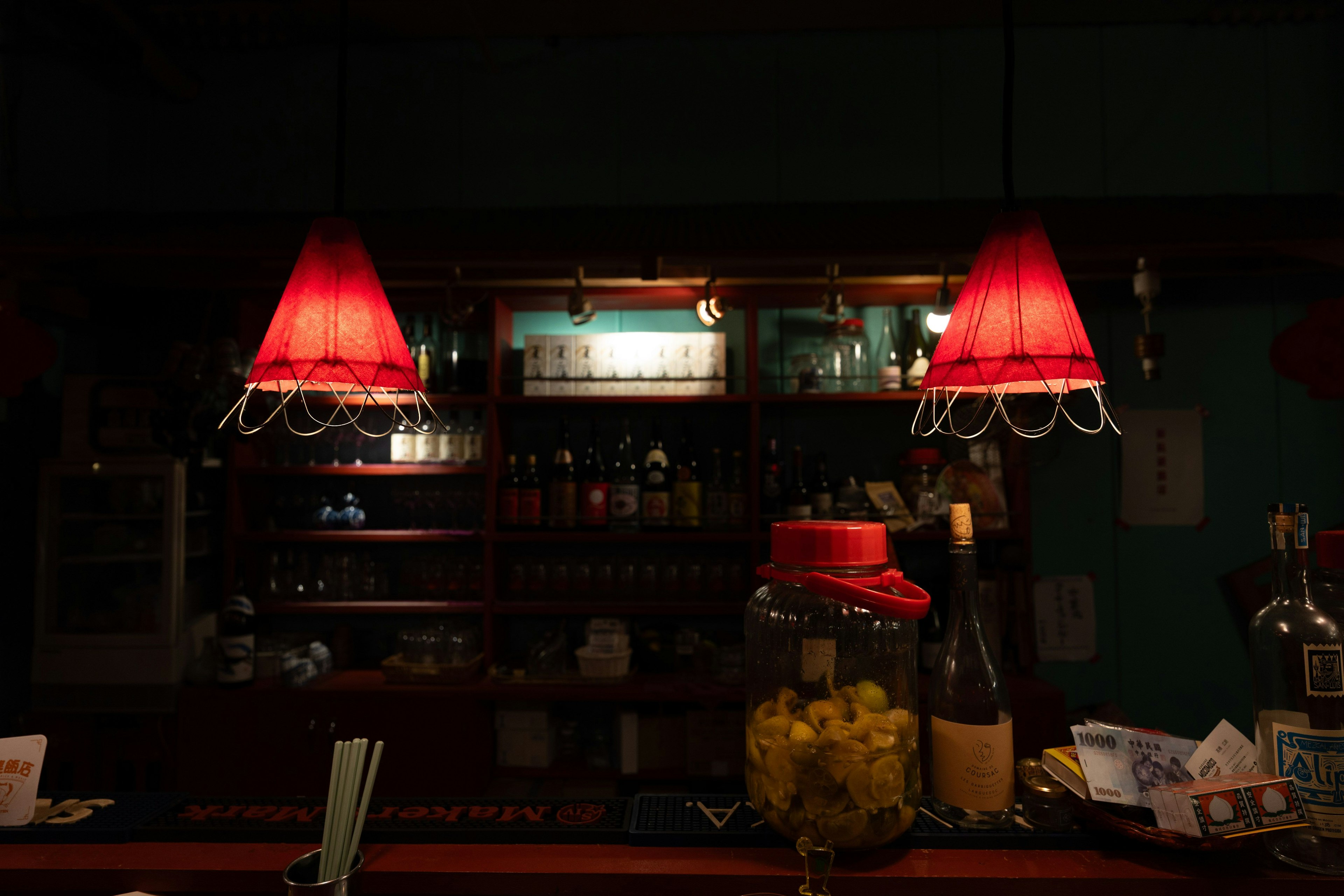 Interior de un bar con pantallas rojas colgando sobre el mostrador botellas y vasos visibles en el fondo oscuro