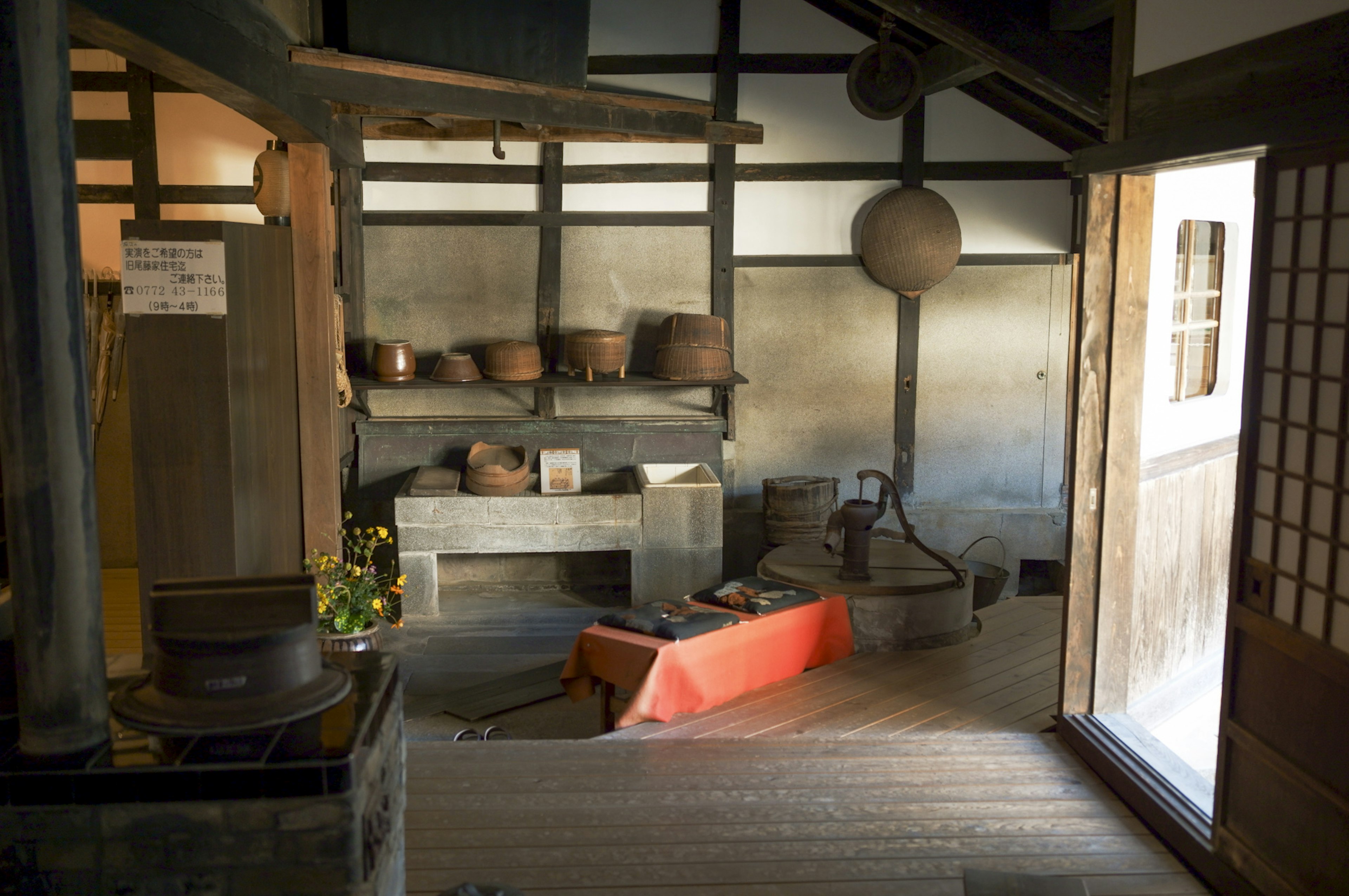 Interior de una casa japonesa tradicional con suelo de madera, suelo de barro, mesa cubierta con tela roja, estantes con ollas de cerámica