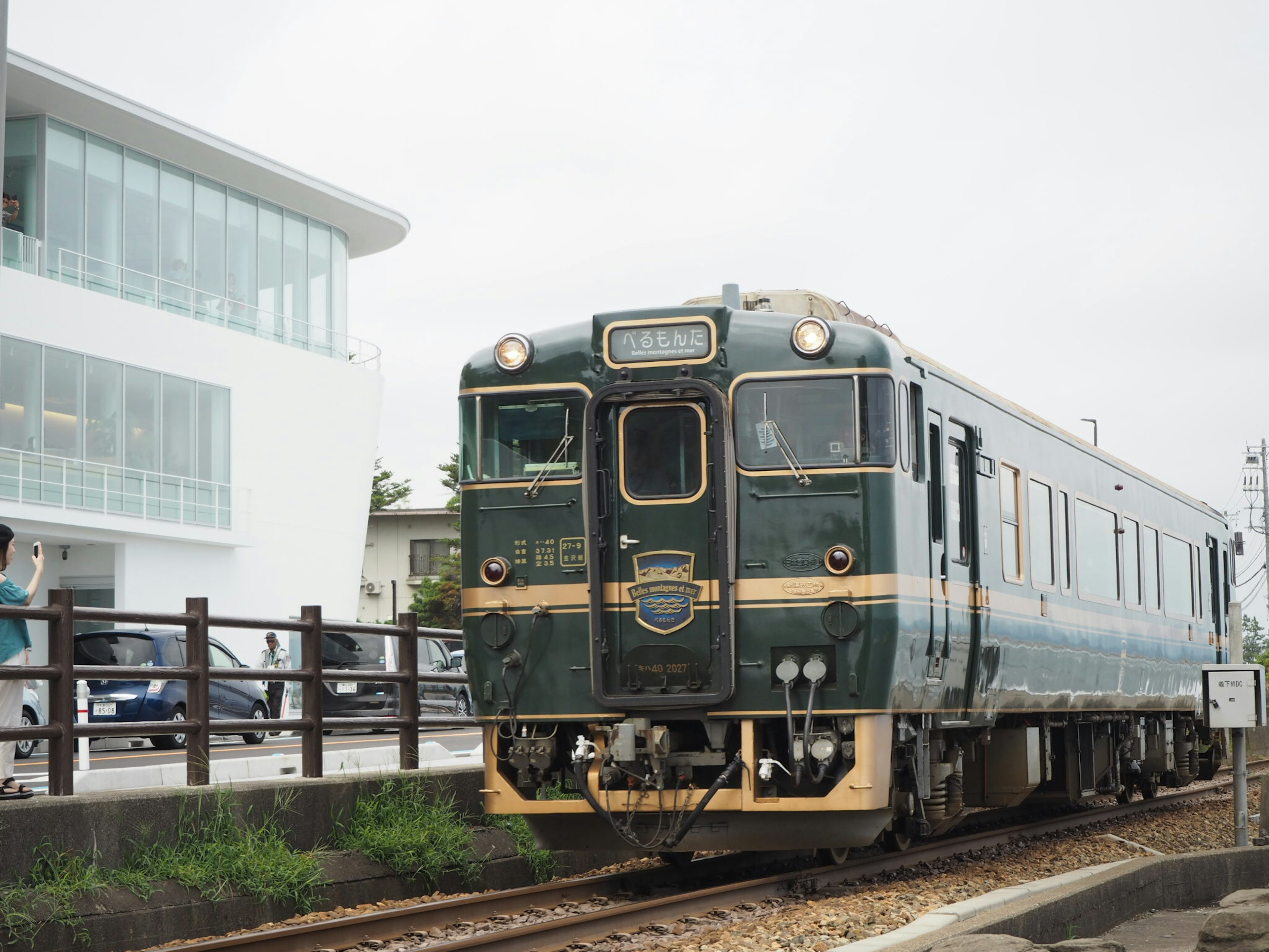 A vintage green train stops at a station with a modern building in the background