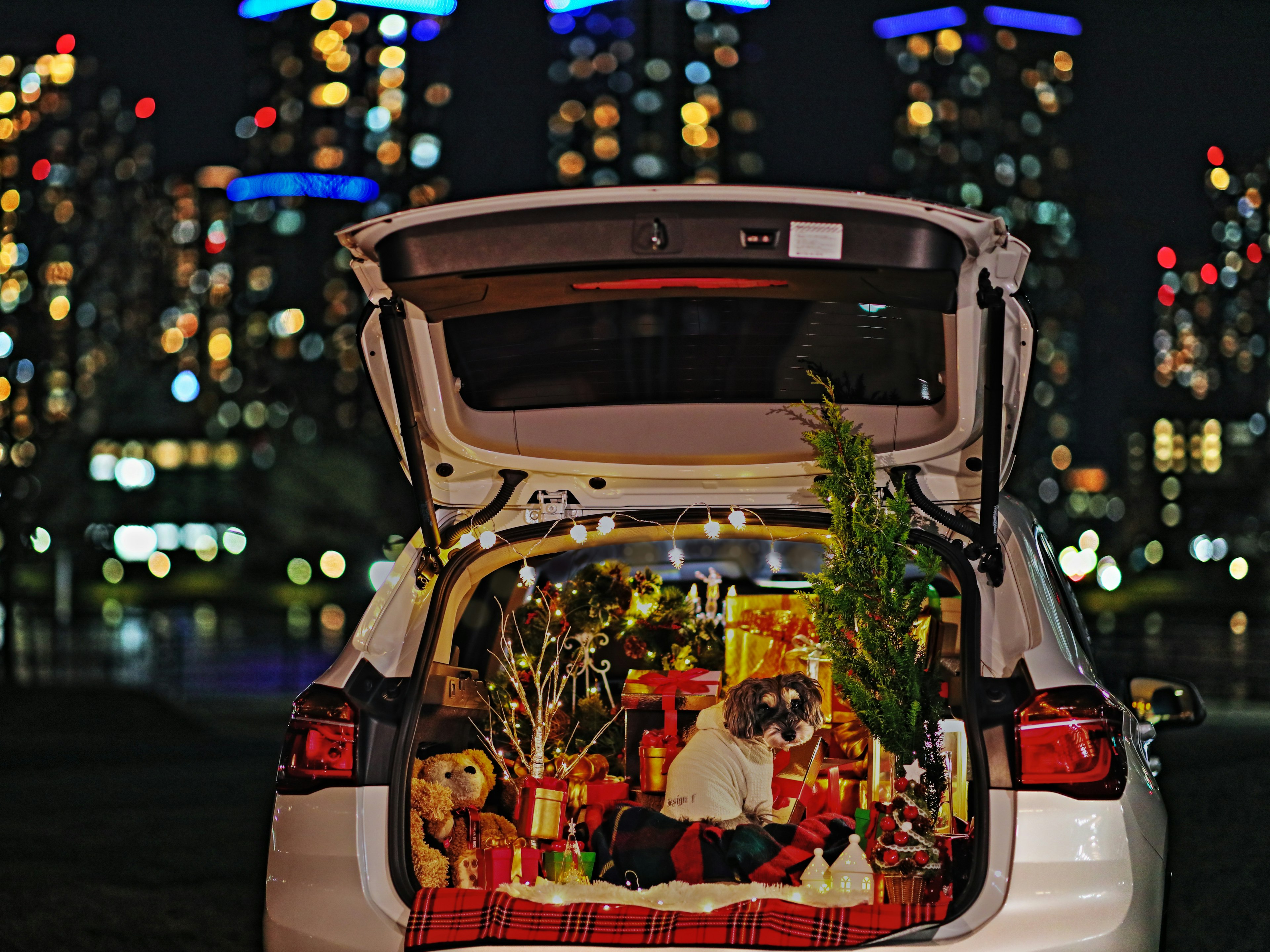 Maletero de coche abierto lleno de regalos y decoraciones navideñas con el horizonte de la ciudad de noche