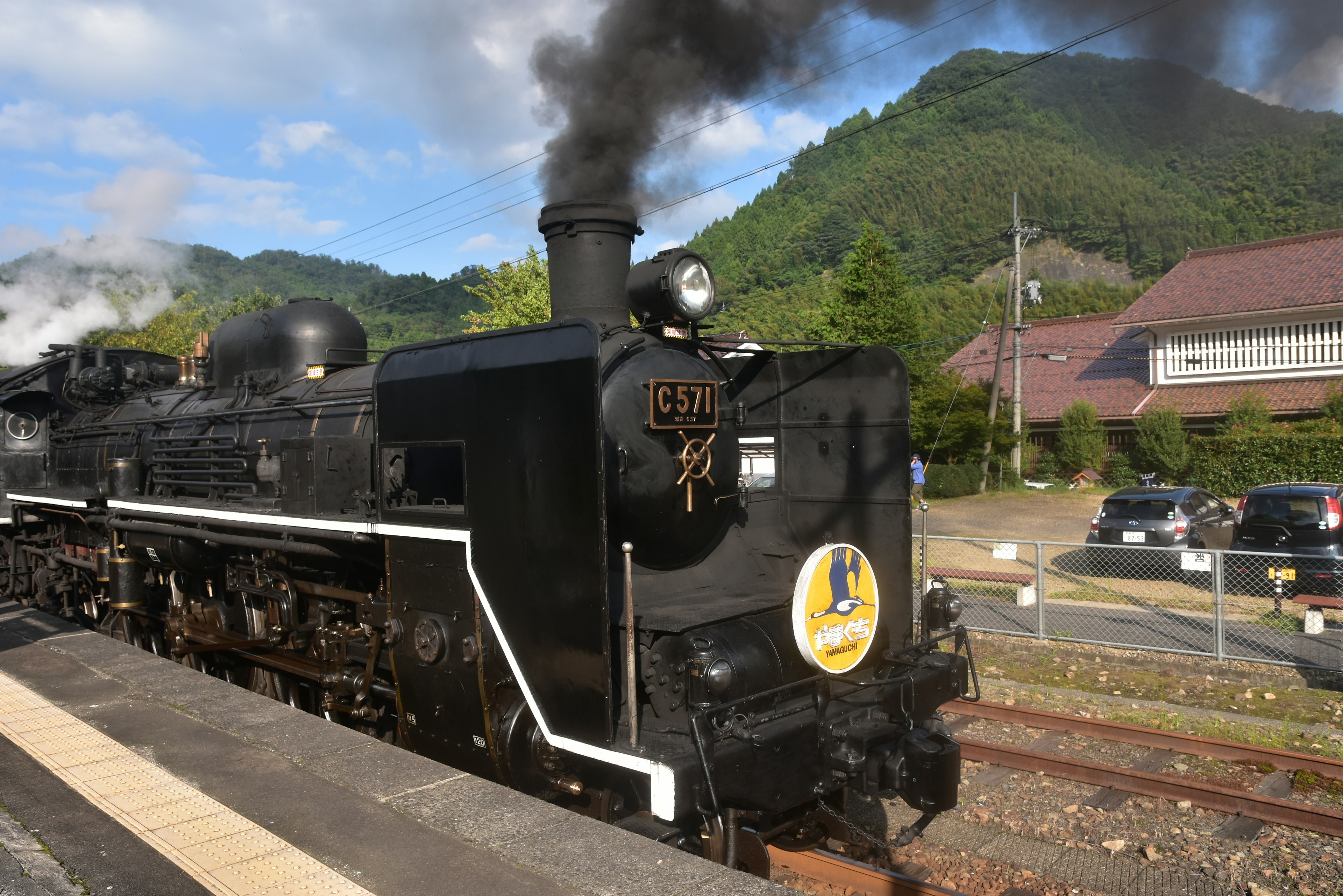 Locomotora de vapor negra emitiendo humo en una plataforma con montañas al fondo