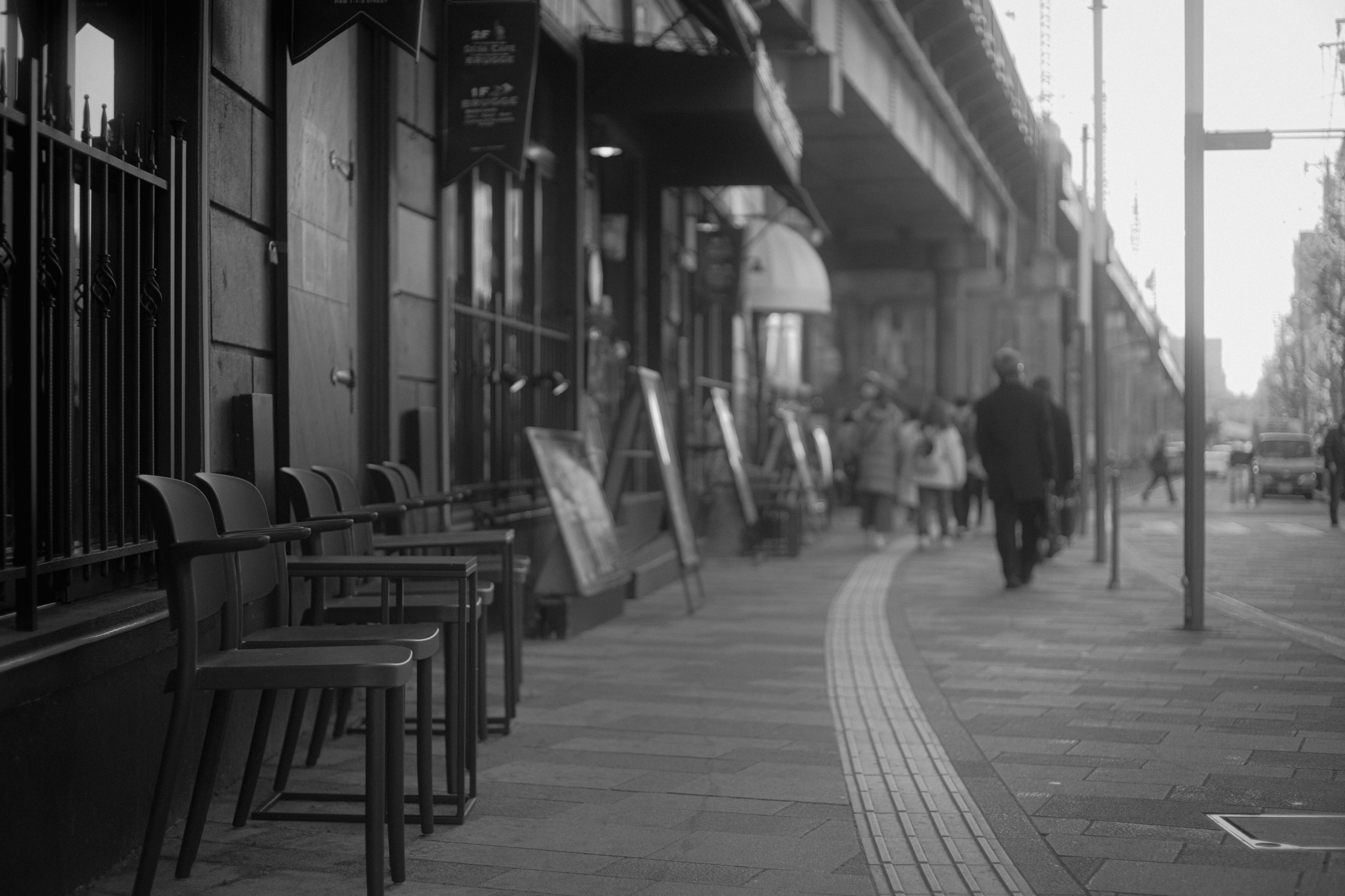 Scène de rue en noir et blanc avec des chaises devant un café des gens marchant sur le trottoir