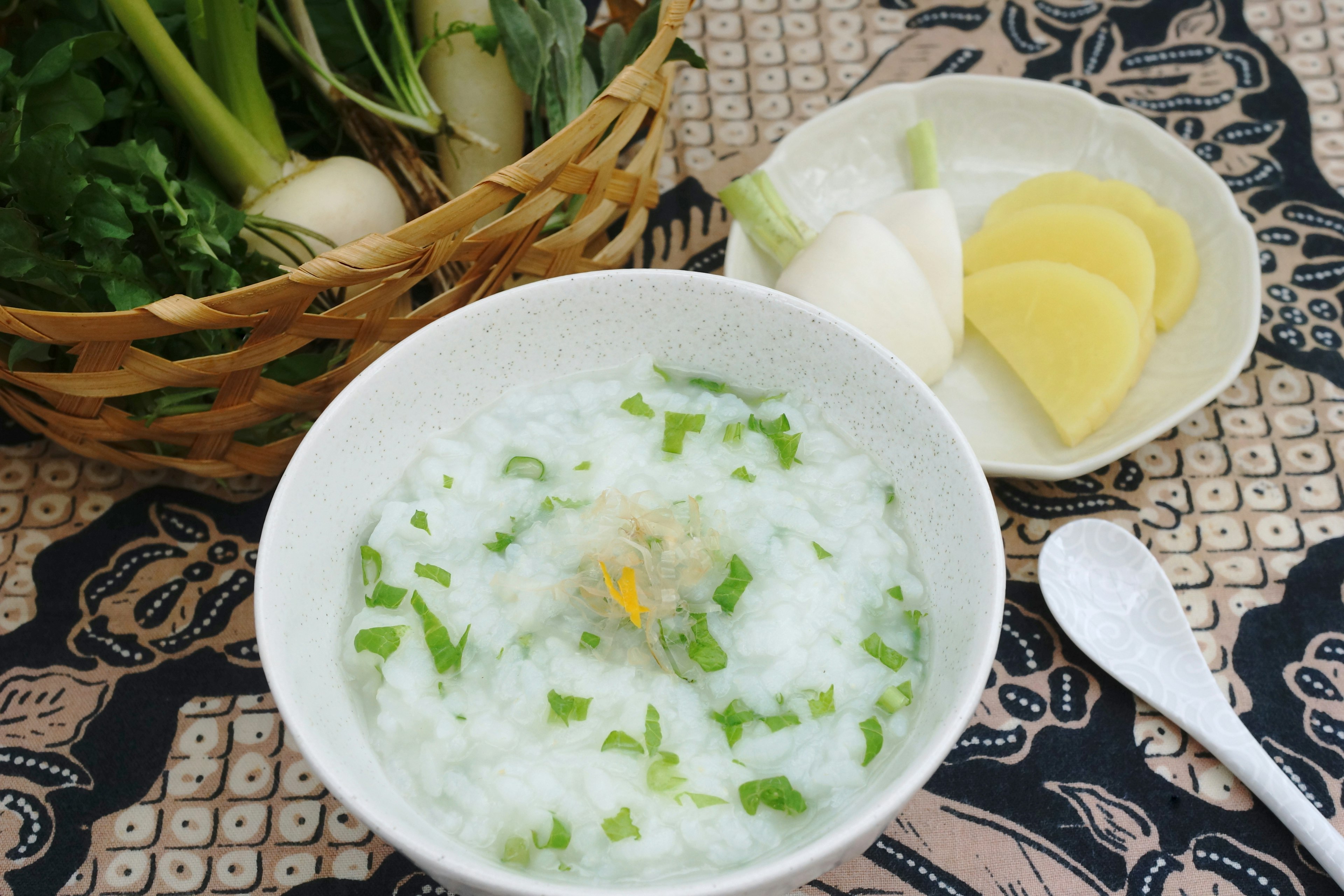 Bowl of white rice porridge topped with green onions and herbs alongside sliced radish and potato