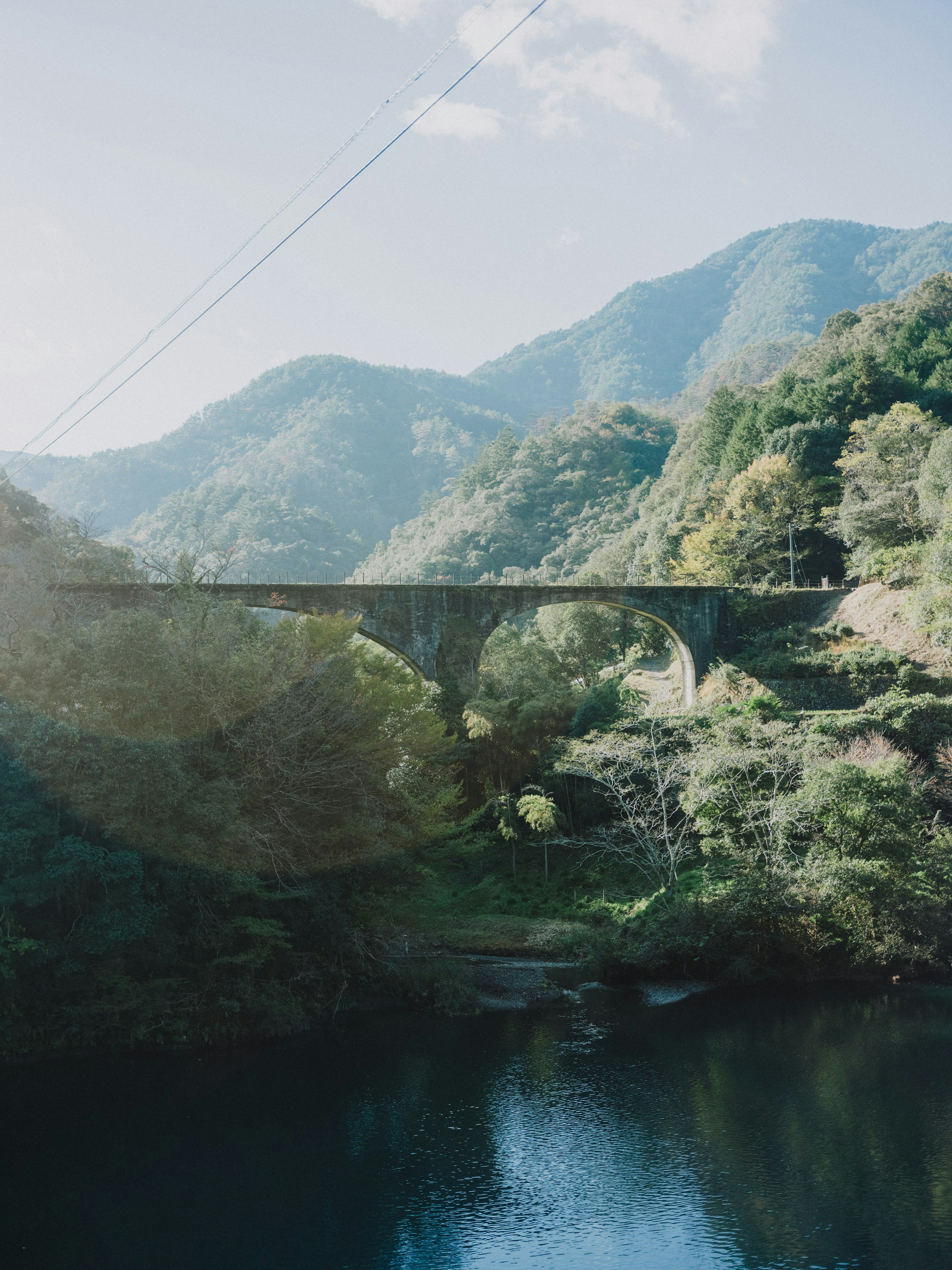 Pemandangan indah jembatan lengkung melintasi sungai dikelilingi pegunungan dan pepohonan hijau