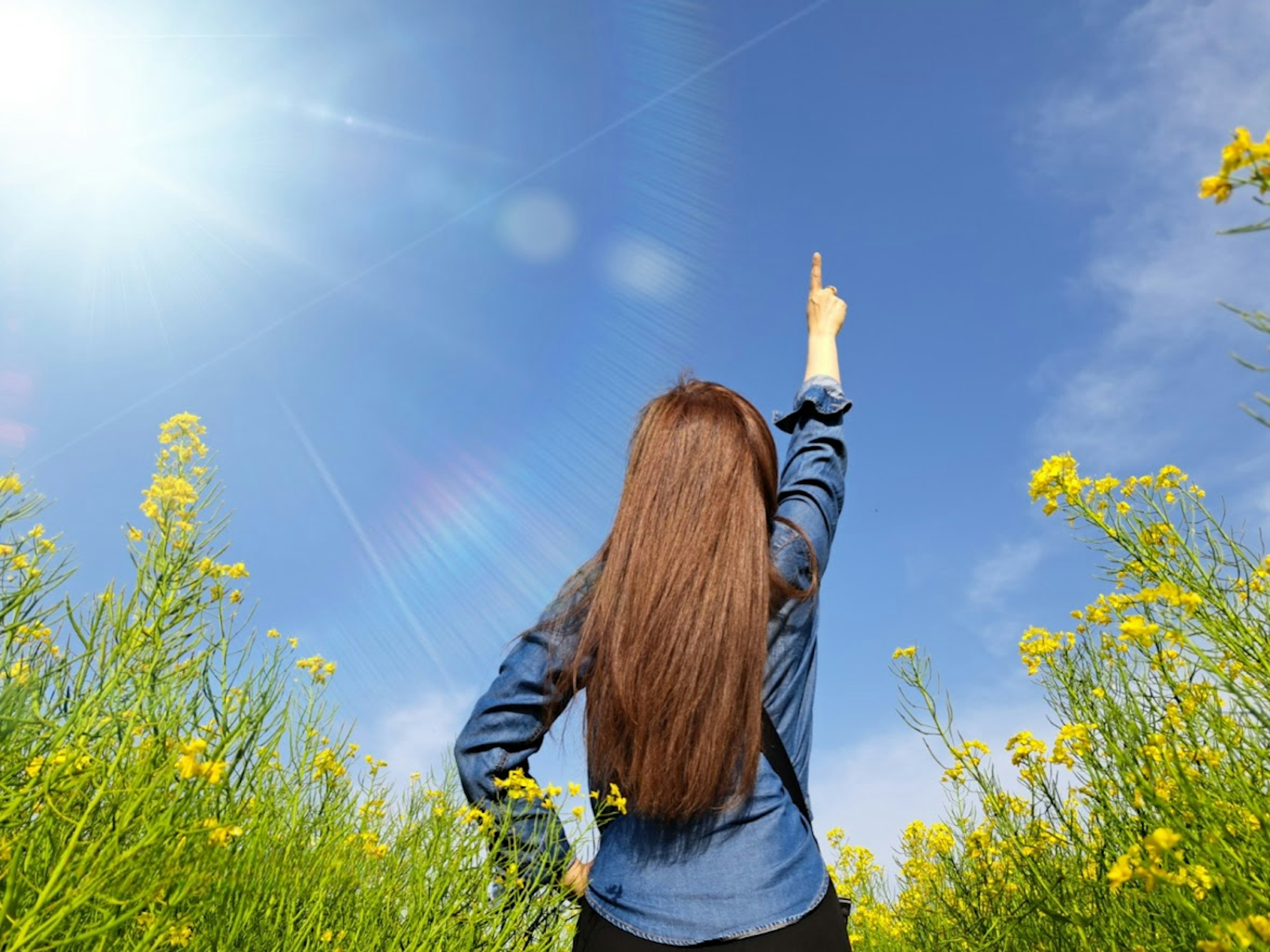 青空の下で黄色い花に囲まれた女性が指を空に向けている