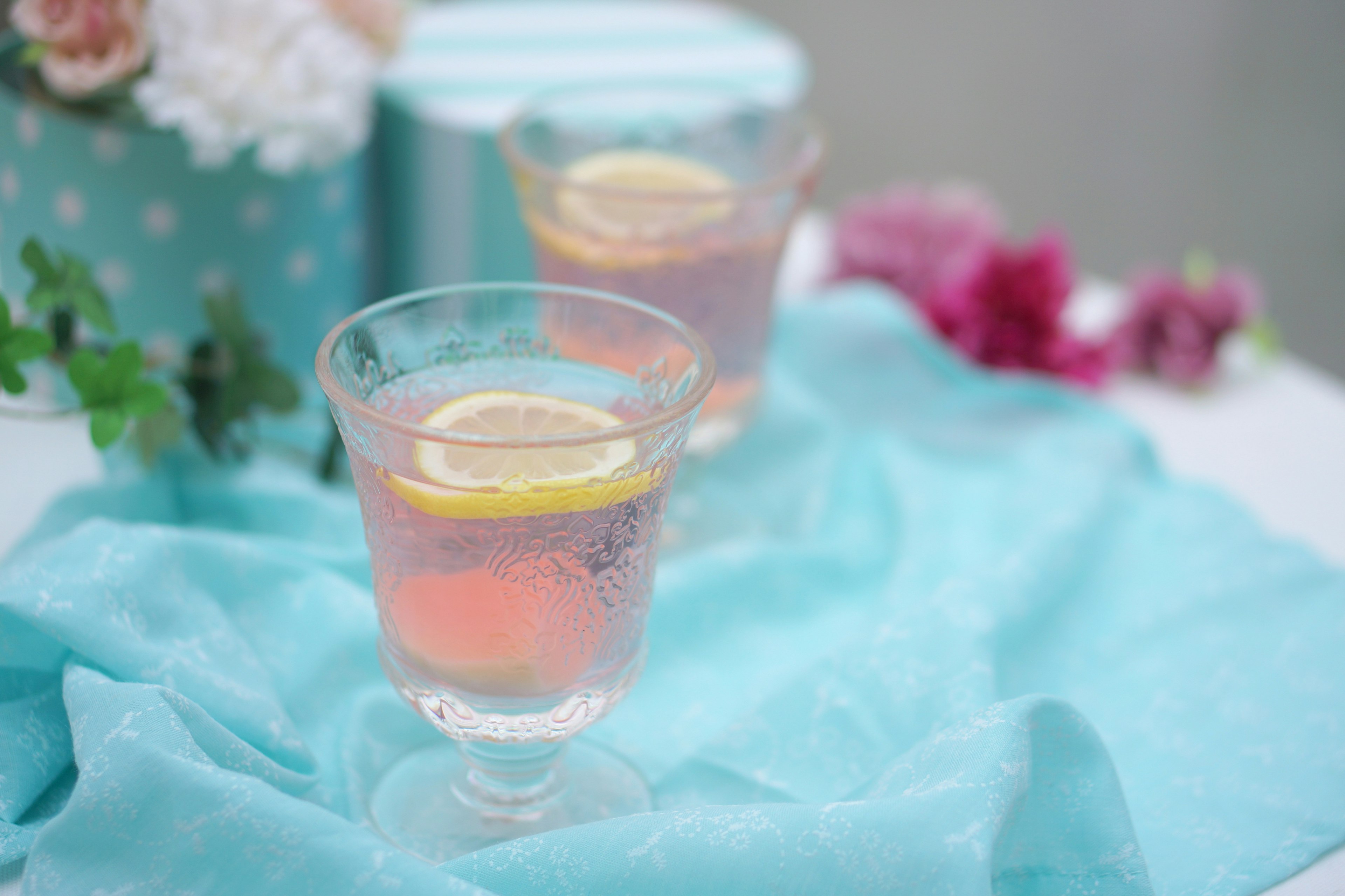 Minuman pink dengan irisan lemon dalam gelas bening di atas kain biru turquoise dengan bunga dekoratif