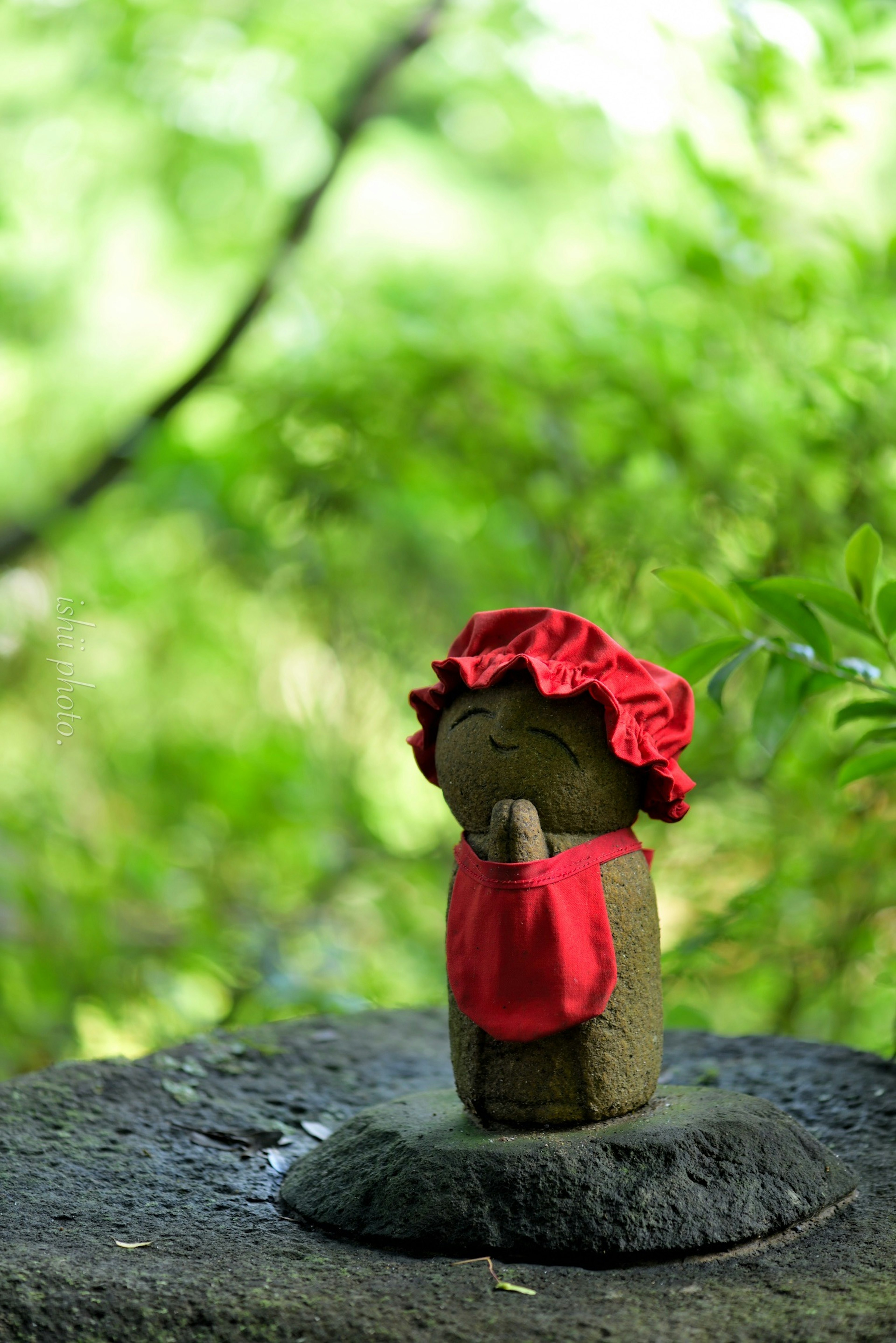 A small stone statue wearing a red hat and apron standing in green nature