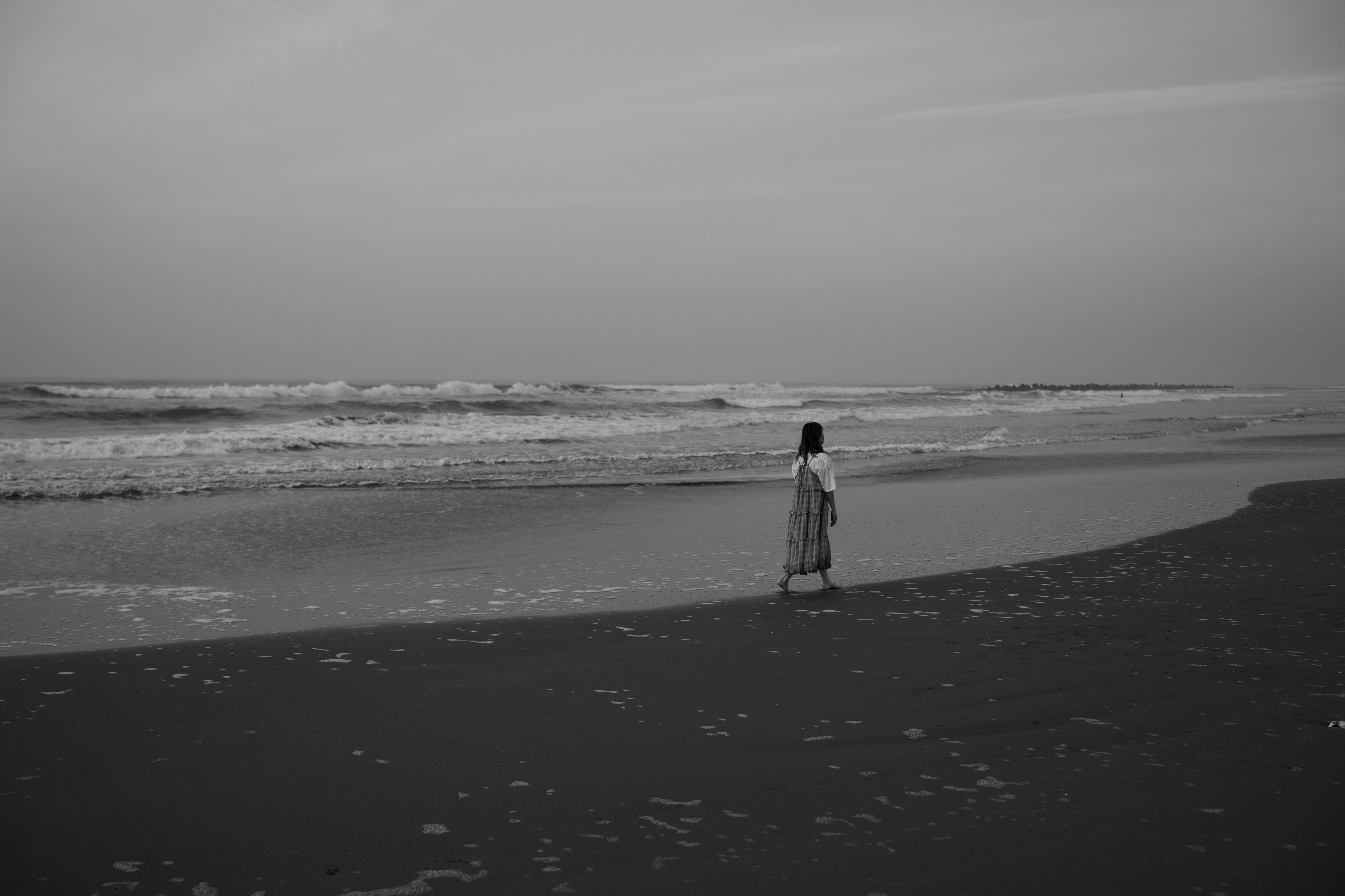 Schwarzweißfoto einer Frau, die am Strand steht und die Wellen beobachtet