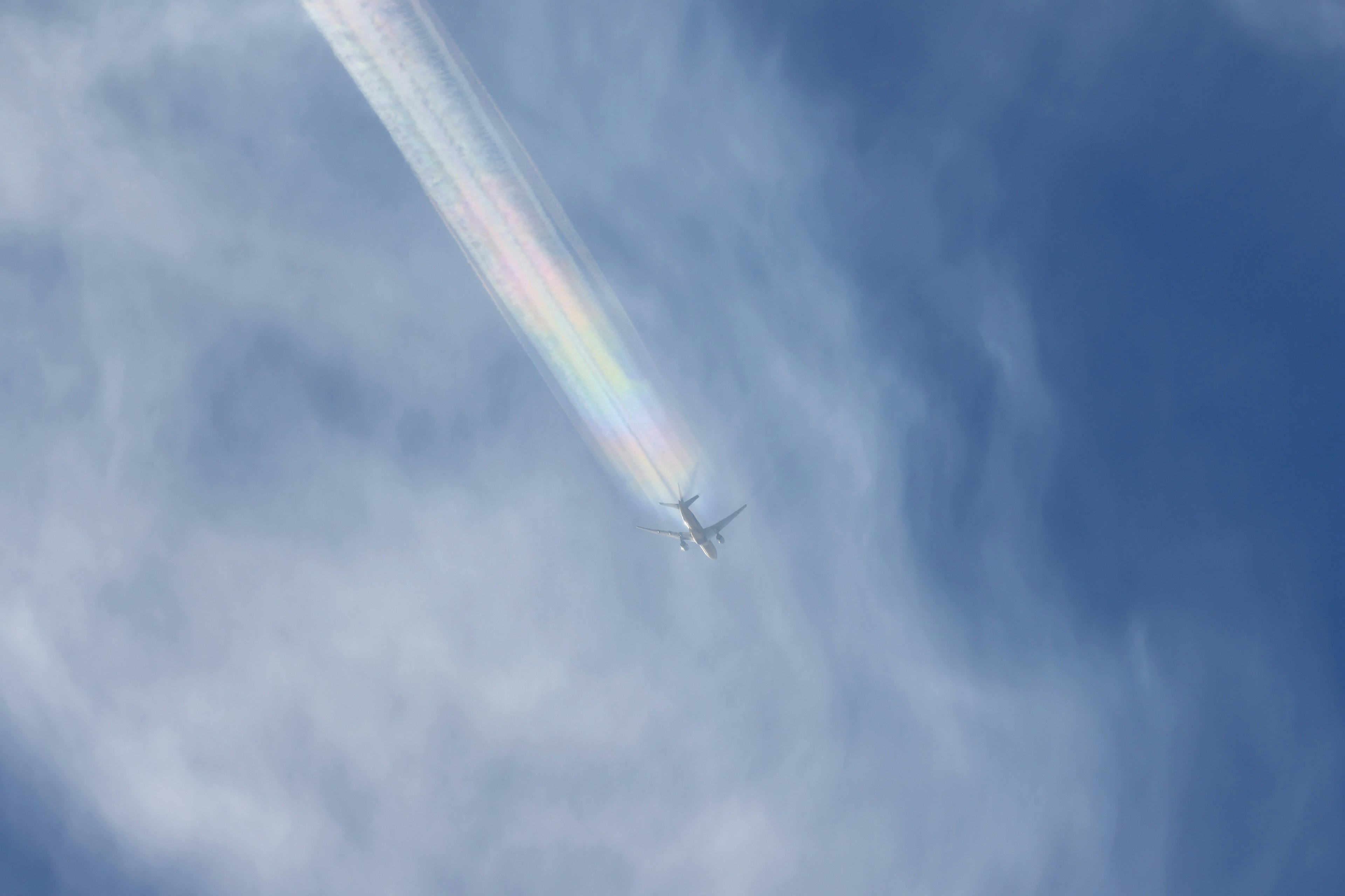 An airplane flying in a blue sky with a rainbow-colored contrail