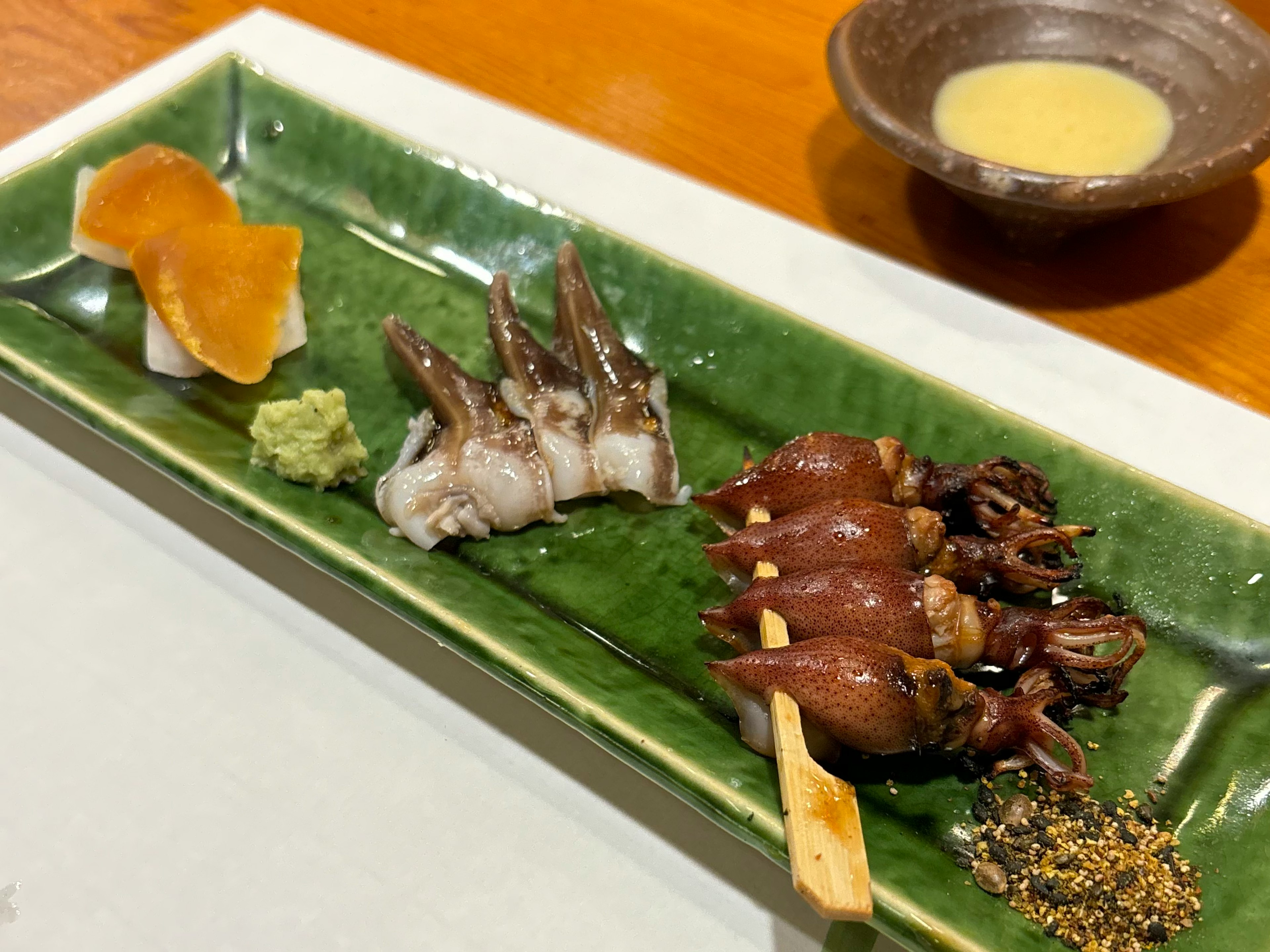 Un plato verde con sashimi y brochetas asadas carne oscura y pescado blanco con wasabi y condimentos al lado