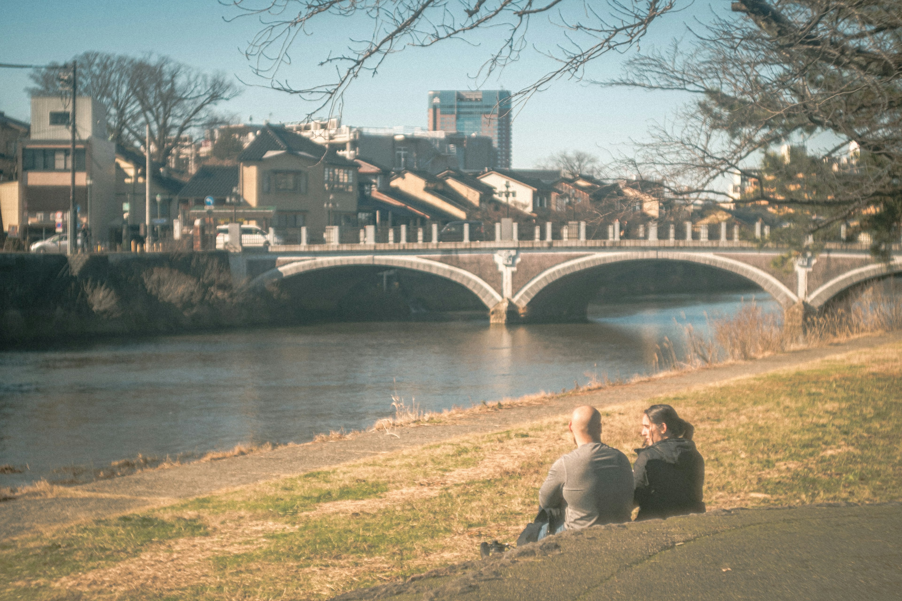 Due persone sedute vicino al fiume con un ponte sullo sfondo