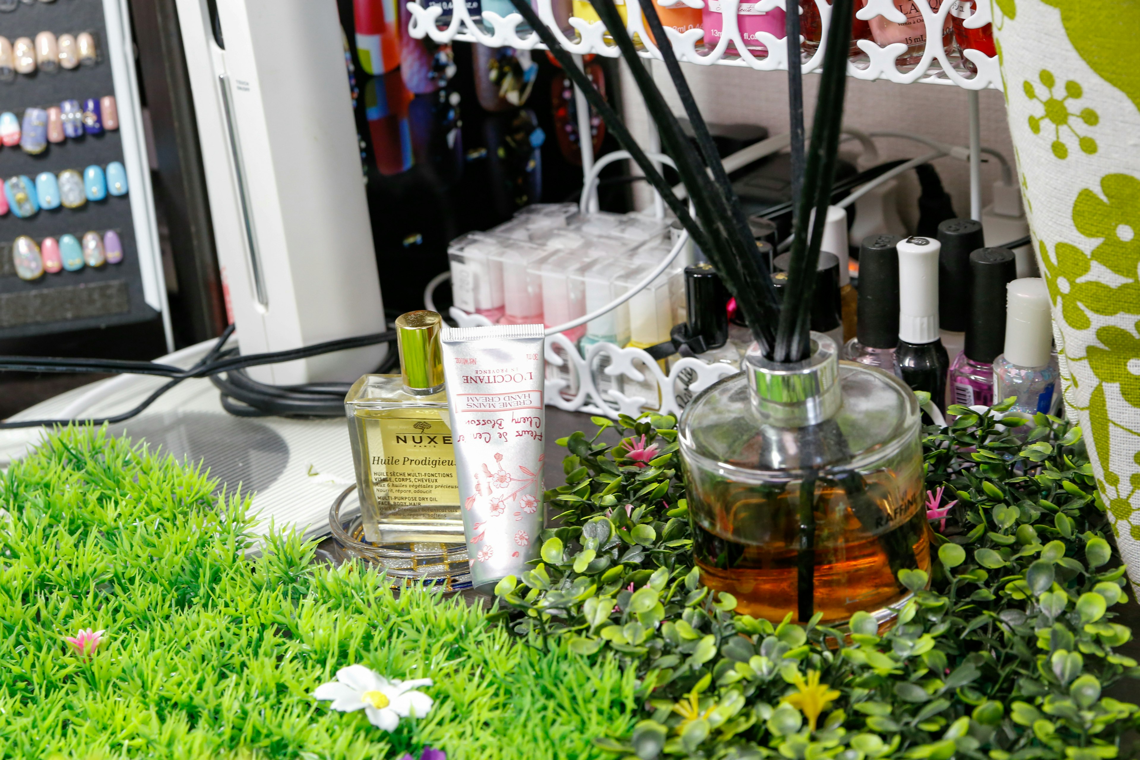 A desktop scene featuring a reed diffuser and cosmetics arranged on green grass