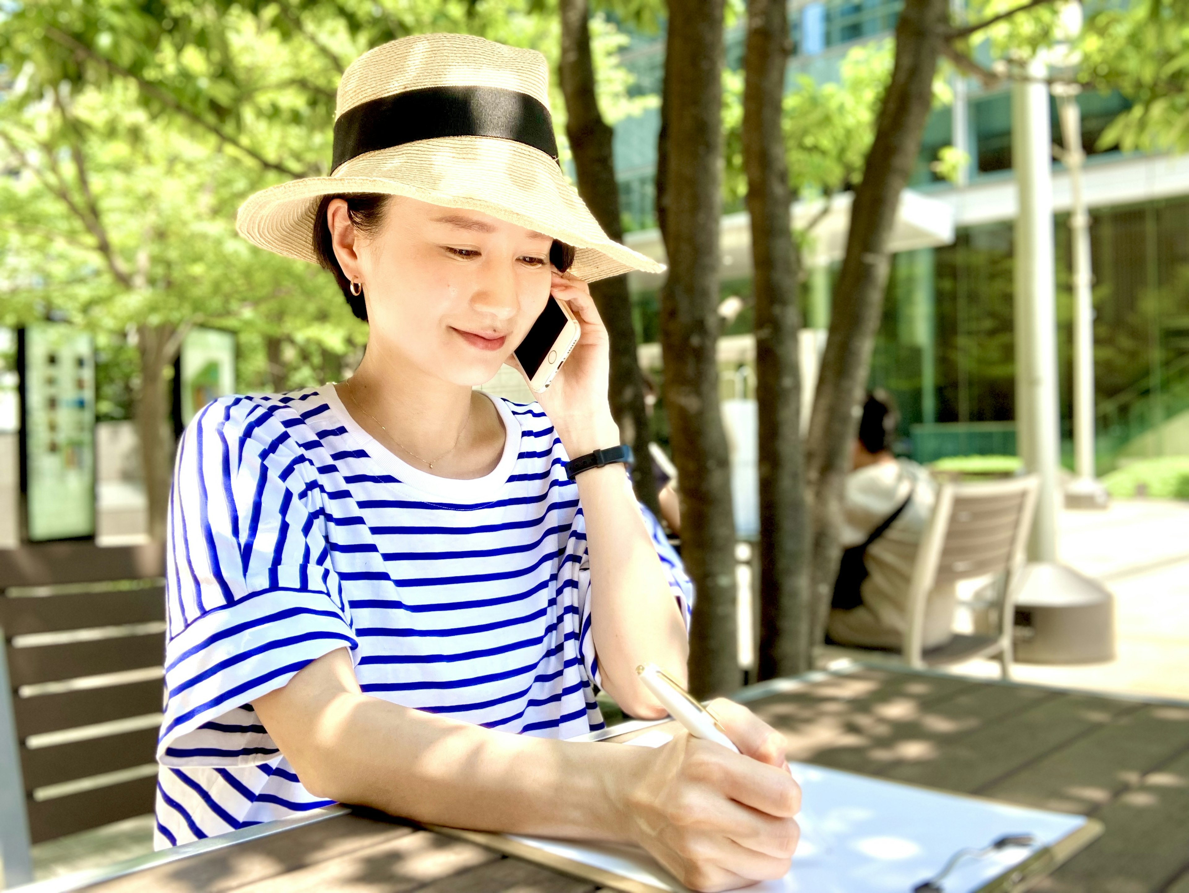 Mujer tomando notas por teléfono con una camisa a rayas y un sombrero café al aire libre con árboles verdes