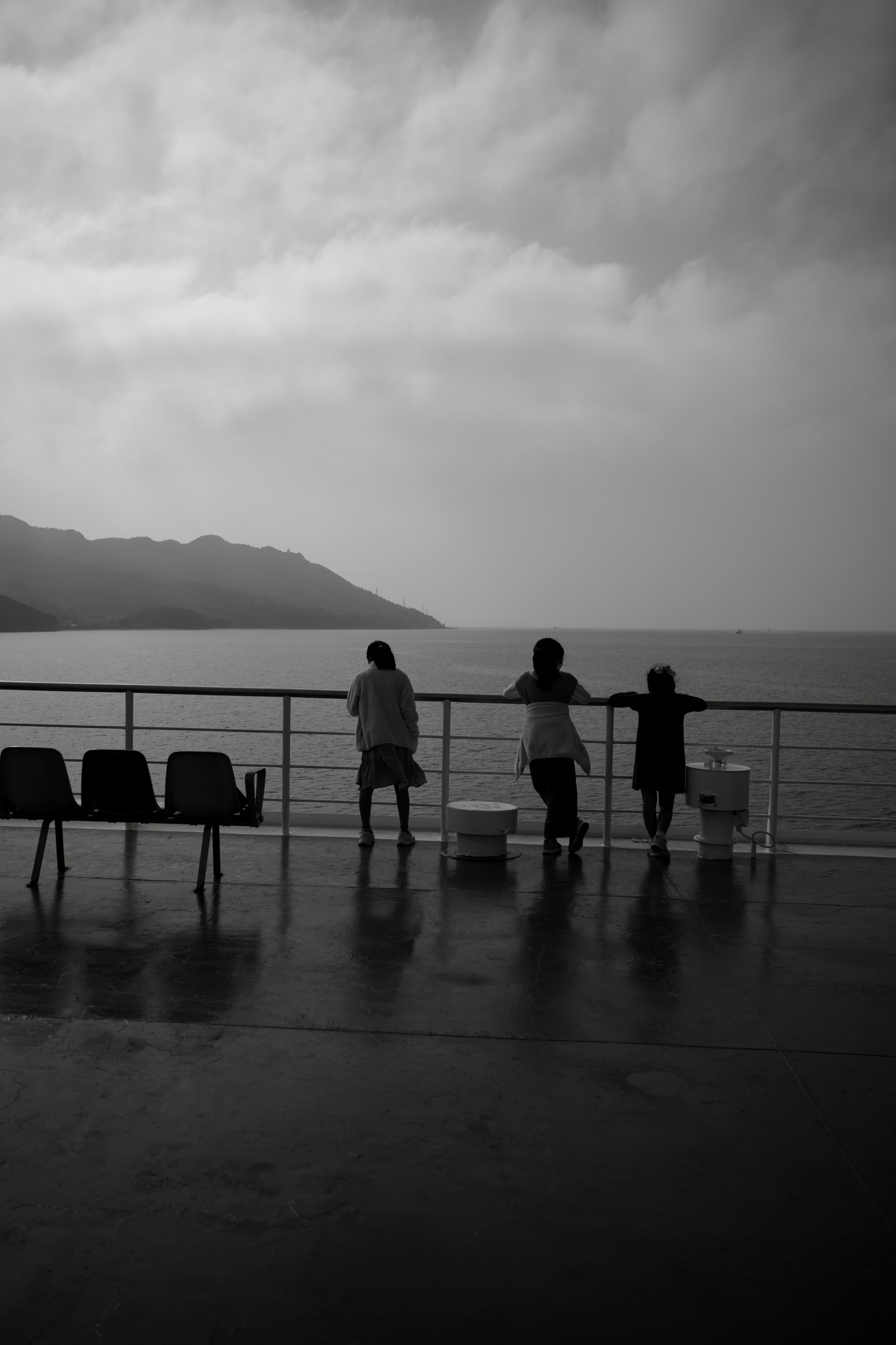 Silhouette von drei Personen, die auf das Meer in einer monochromen Landschaft mit aufgereihten Stühlen schauen