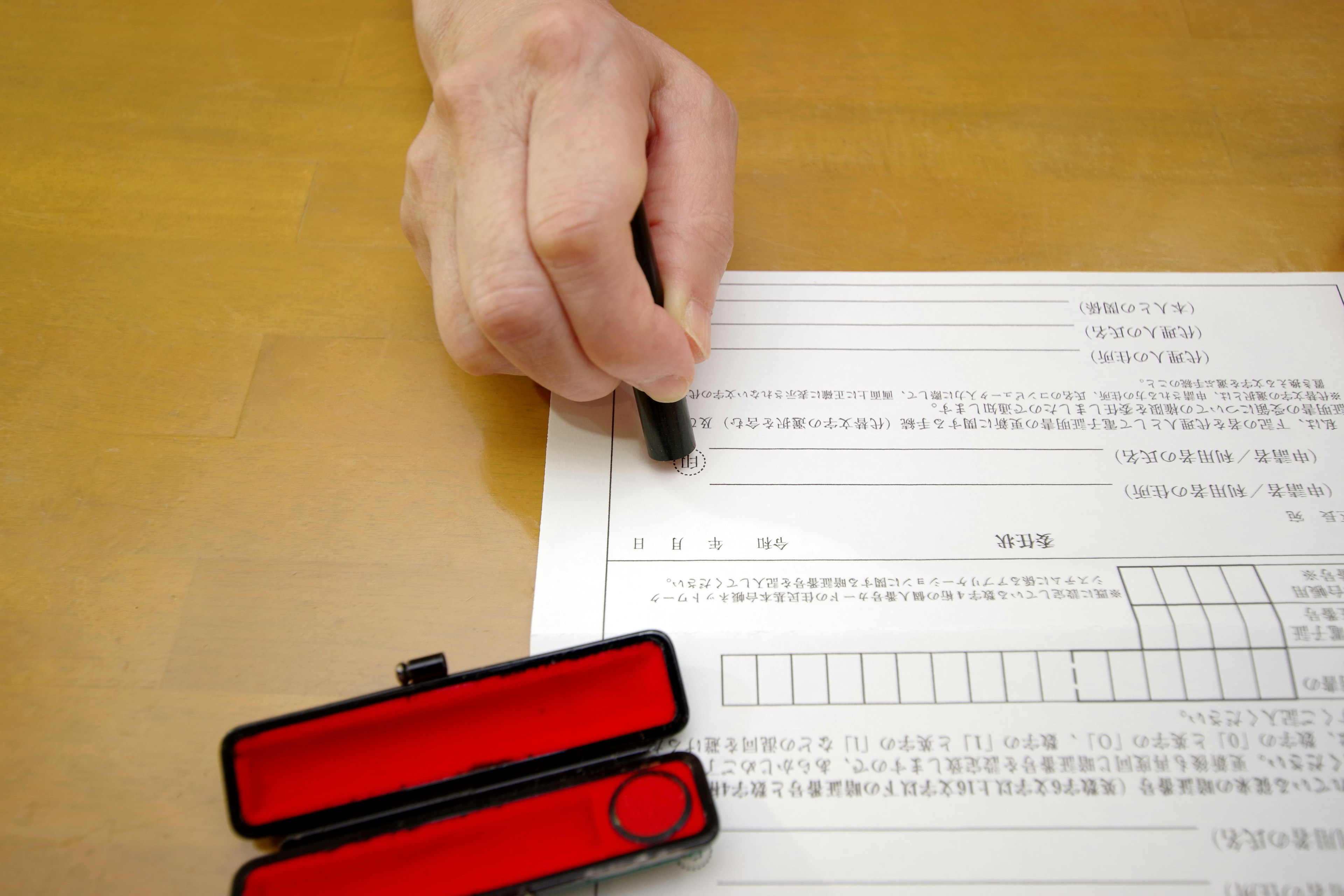 A hand holding a black pen signing a document with a red stamp case beside it