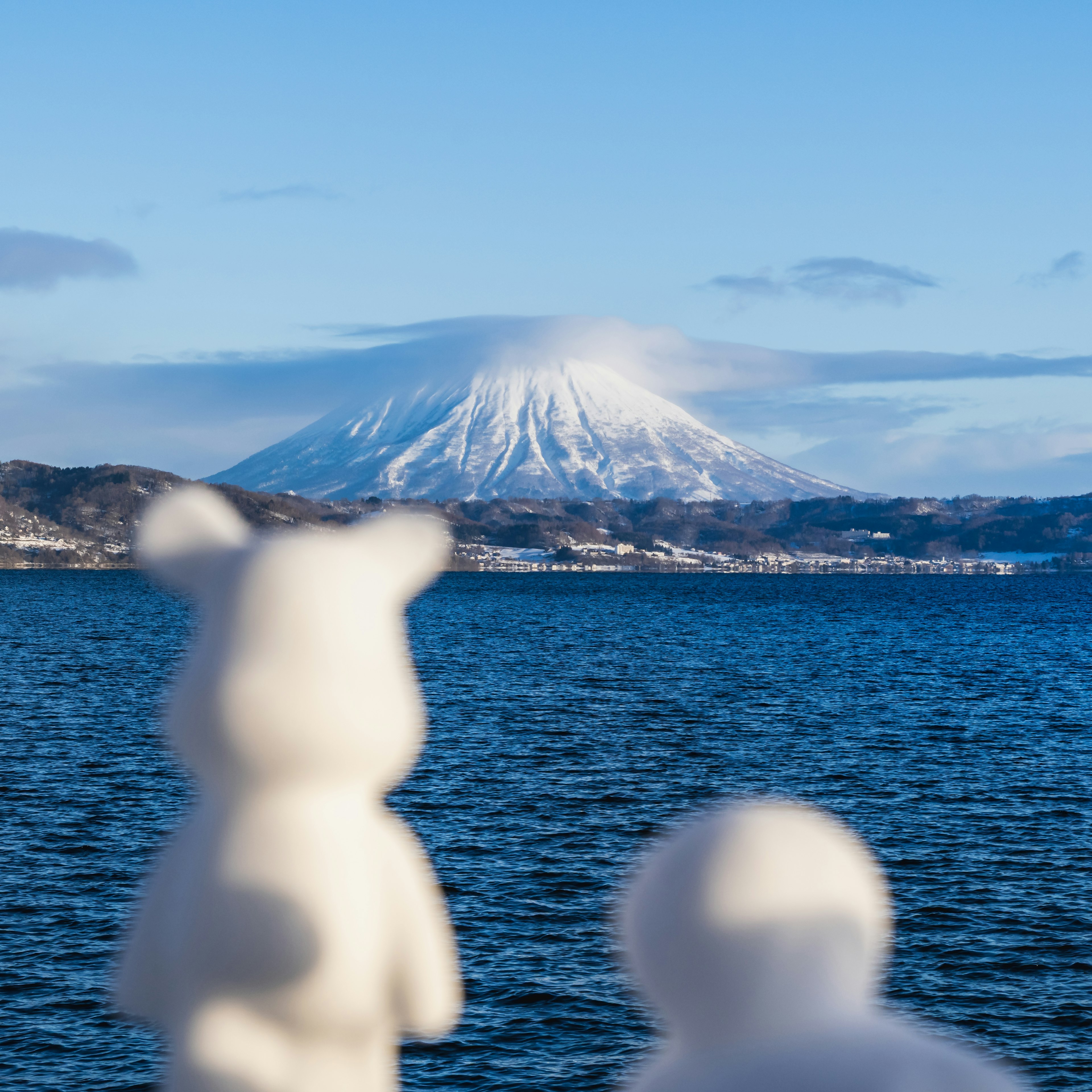 二体の白いクマのフィギュアが海を見つめる背景に雪を被った山がある