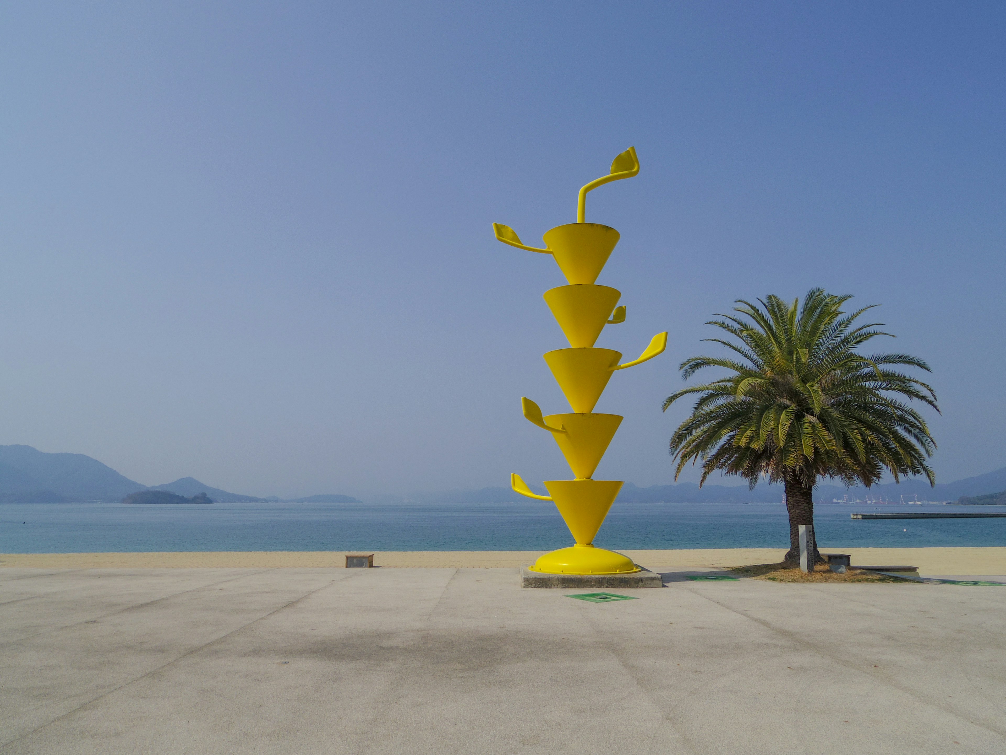 A vibrant yellow sculpture next to a palm tree by the sea