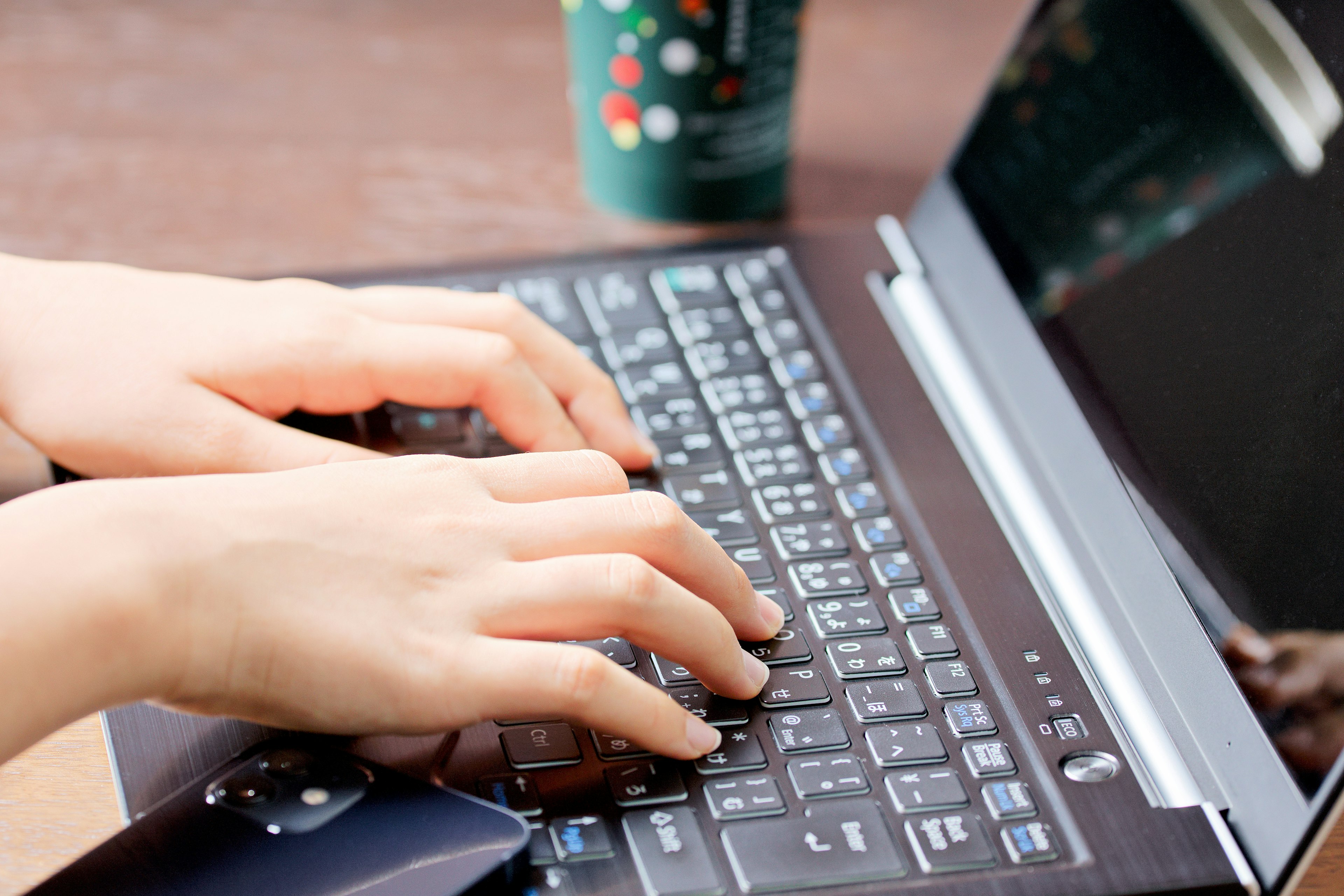 Manos escribiendo en el teclado de una laptop con una taza de café al fondo