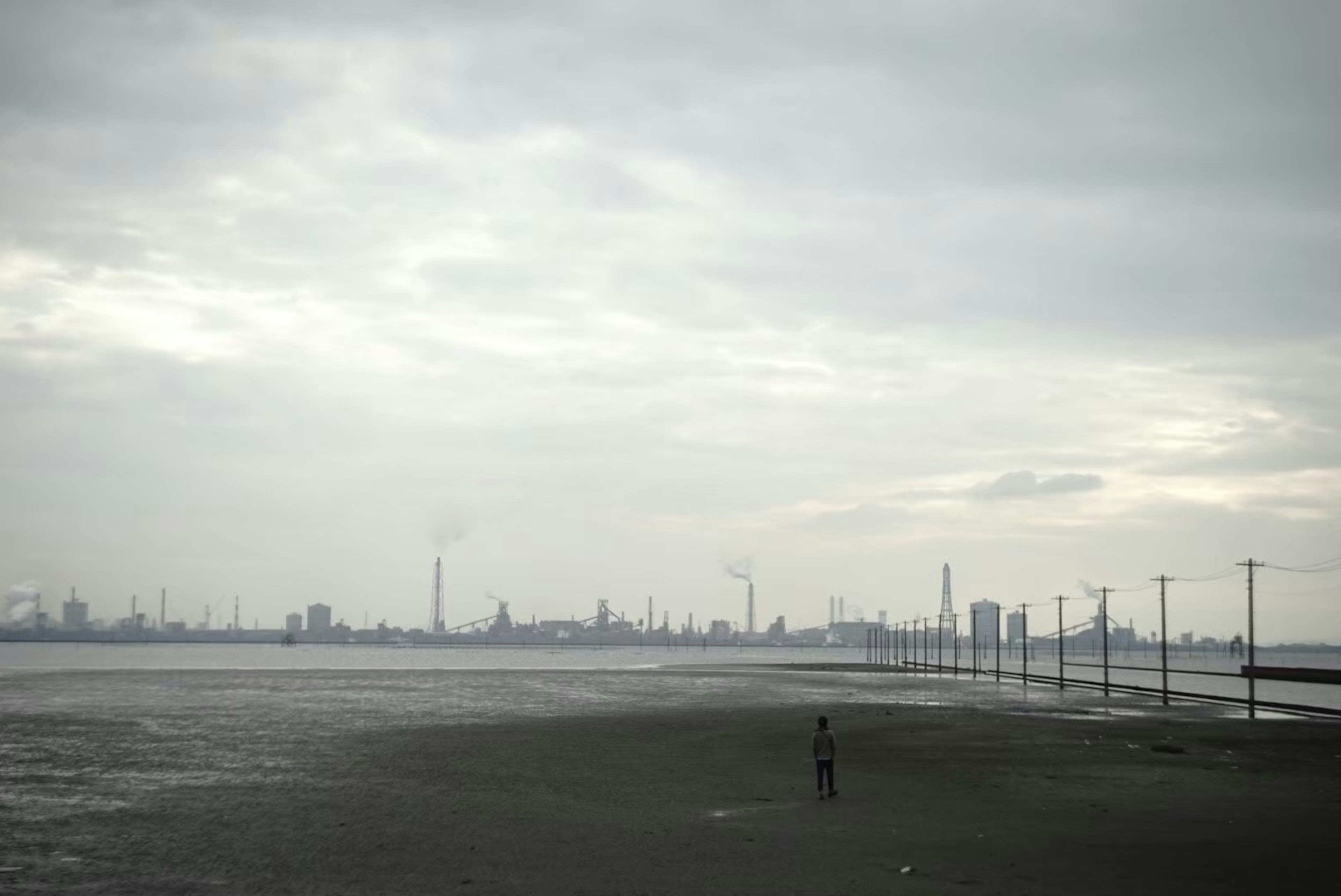 Eine einsame Figur steht an einem Strand unter einem bewölkten Himmel mit einer industriellen Skyline und Stromleitungen im Hintergrund