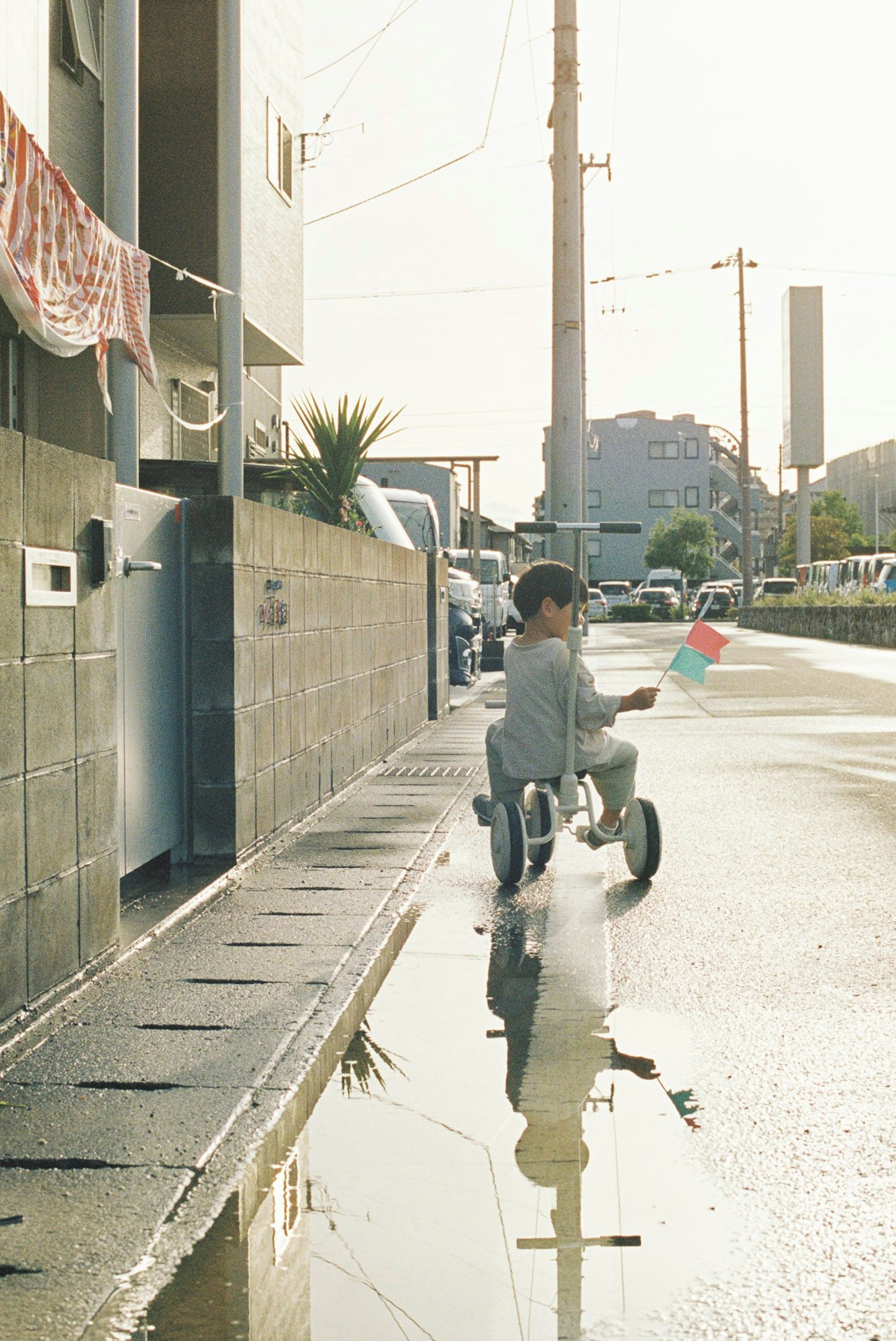 Un niño montando un triciclo en una calle con reflejos en un charco y edificios al fondo