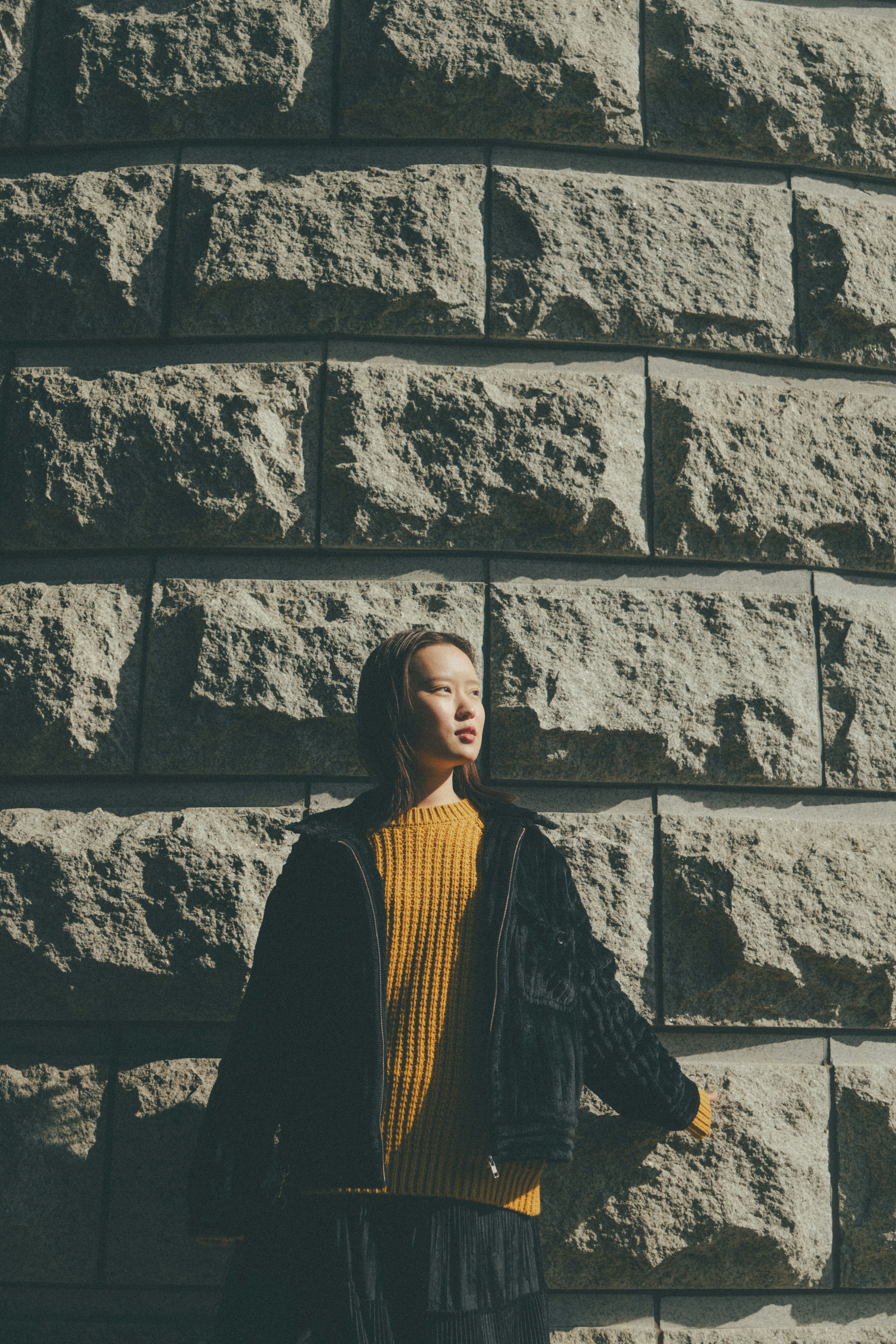 Seorang wanita berdiri di depan dinding batu mengenakan sweter kuning cerah dan jaket hitam