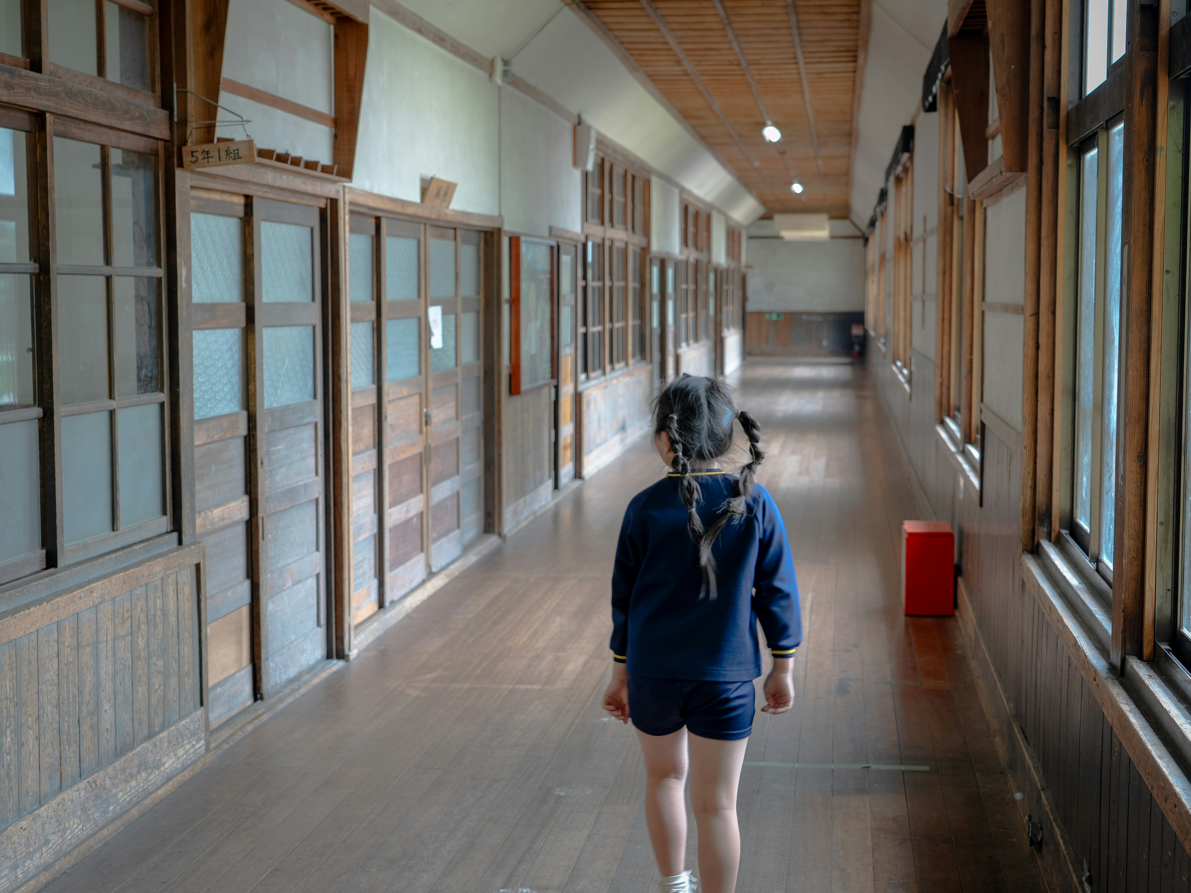 A girl walking down an old school hallway with wooden floors and large windows