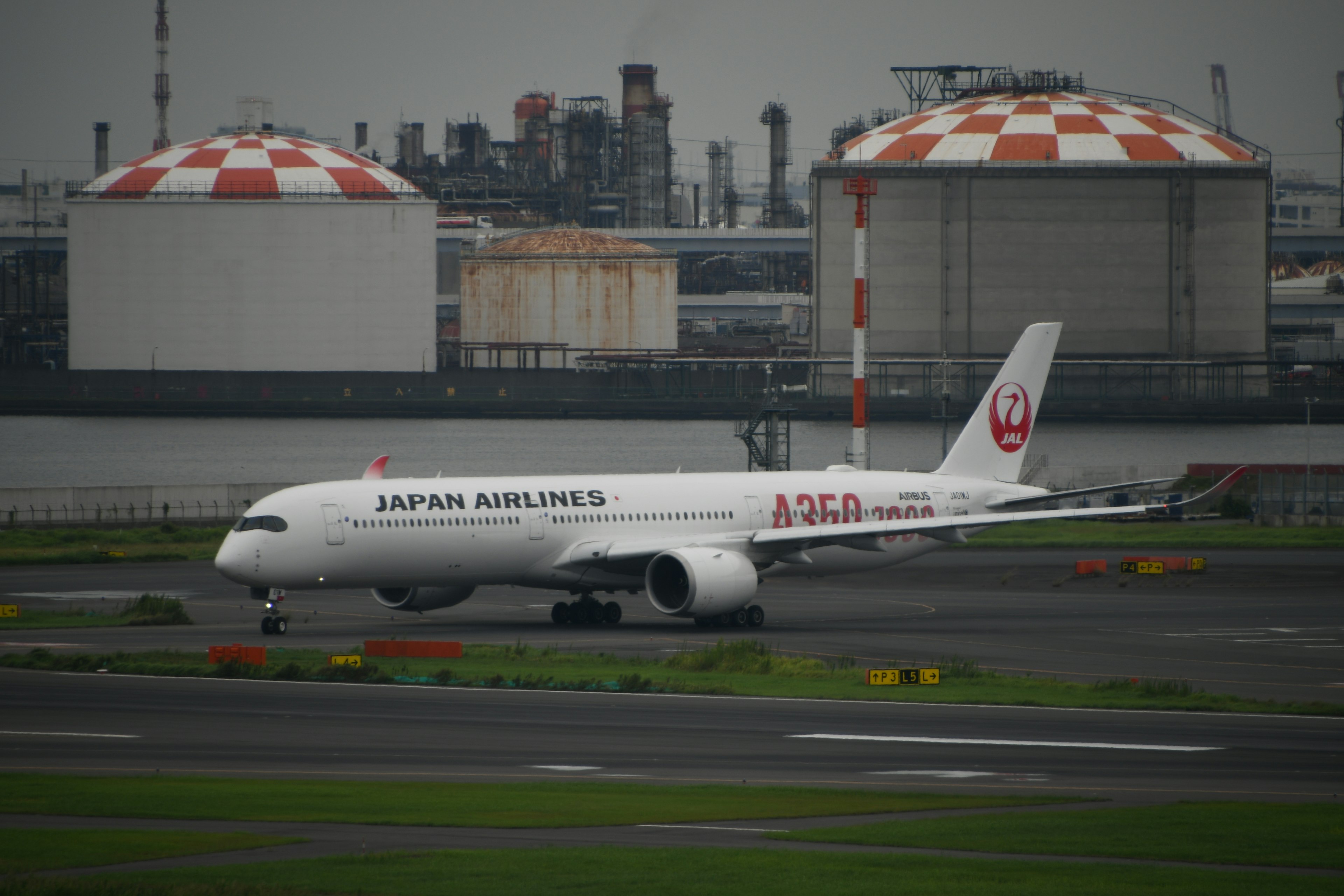 日本航空の飛行機が滑走路を移動している背景に赤と白のタンクが見える