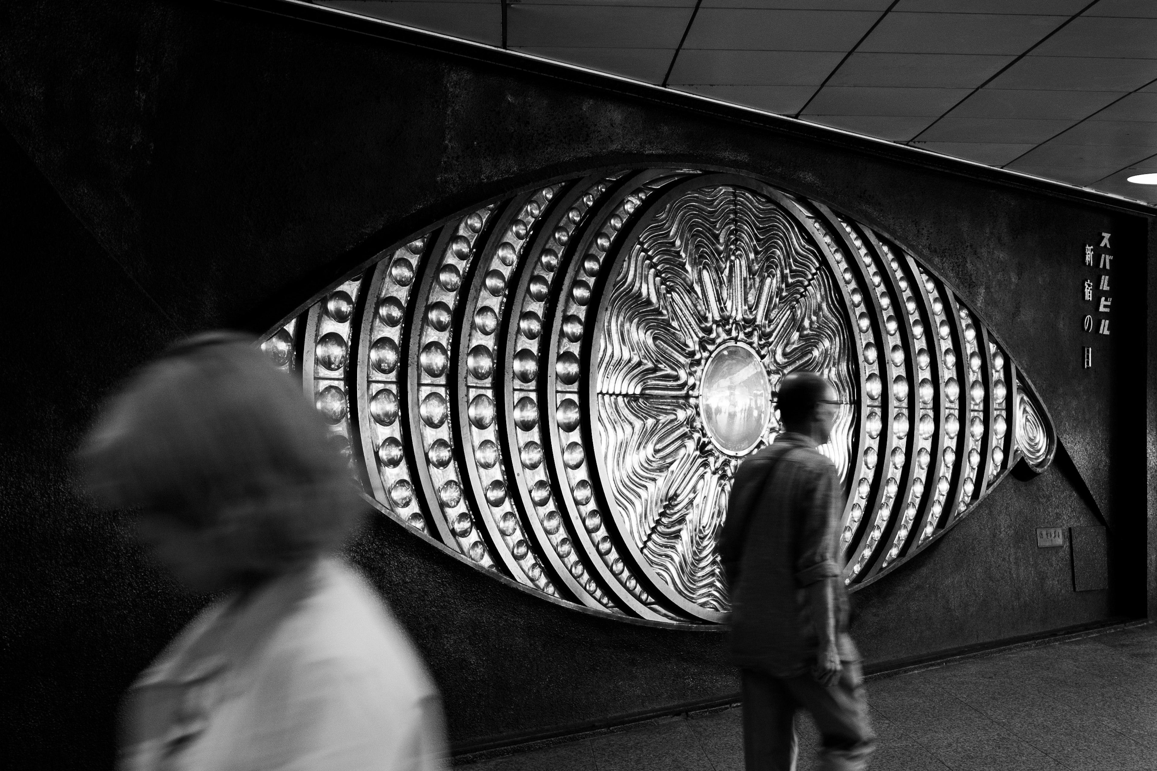 Installation artistique d'un grand œil sur un mur noir avec des piétons passant