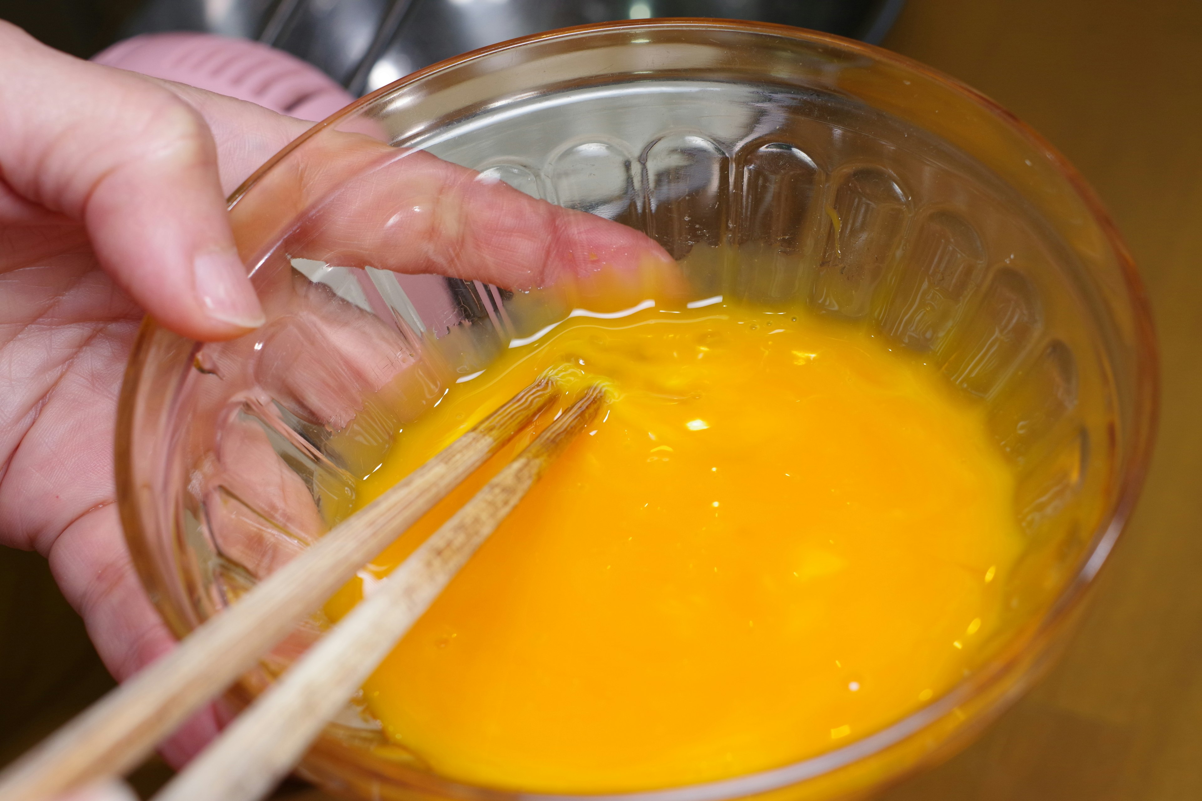 Une main mélangeant des œufs avec des baguettes dans un bol transparent