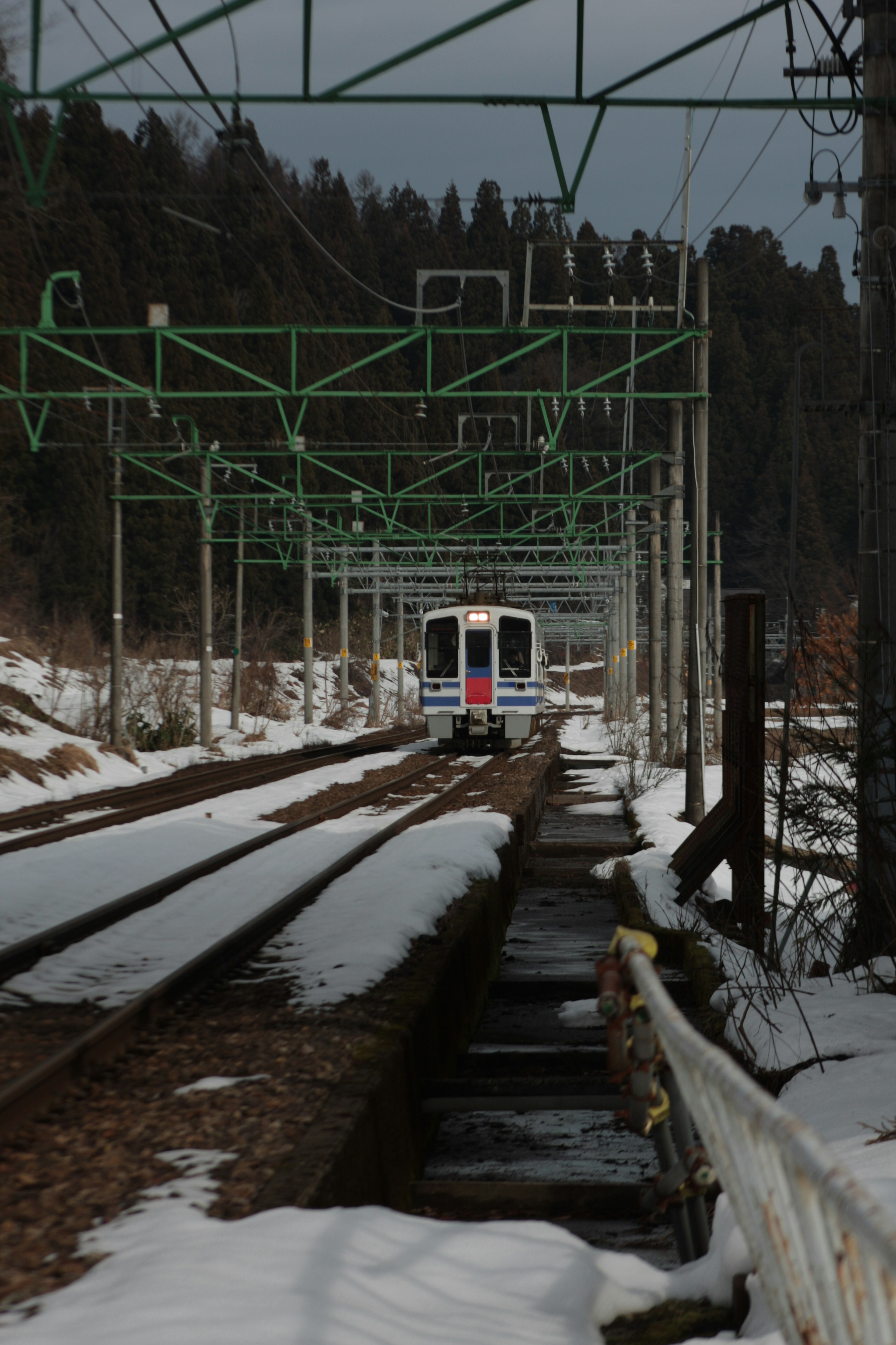 Train on snow-covered tracks with green overhead lines in a rural setting