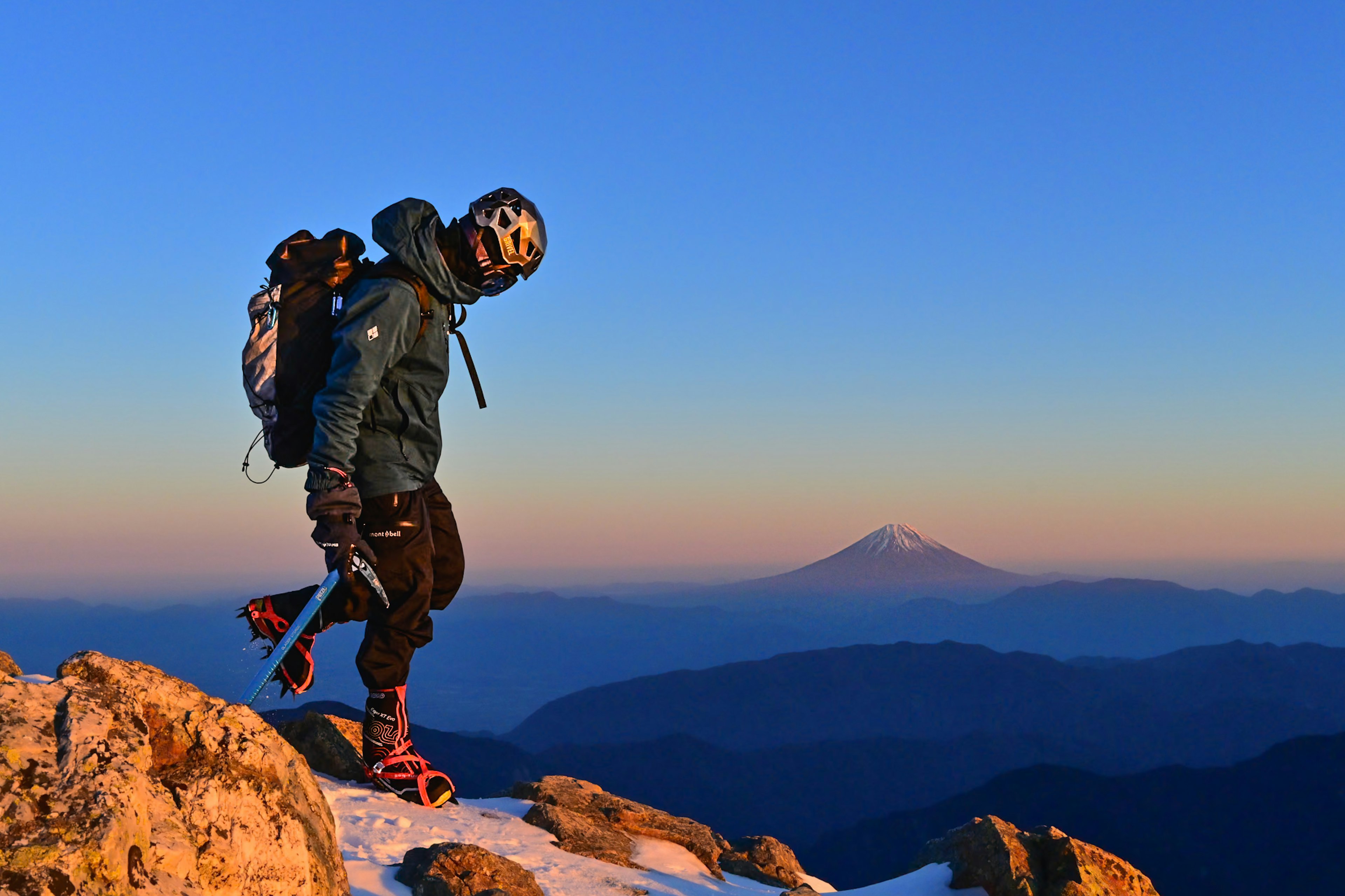 A climber walking on a mountain peak with a snow-capped mountain in the background