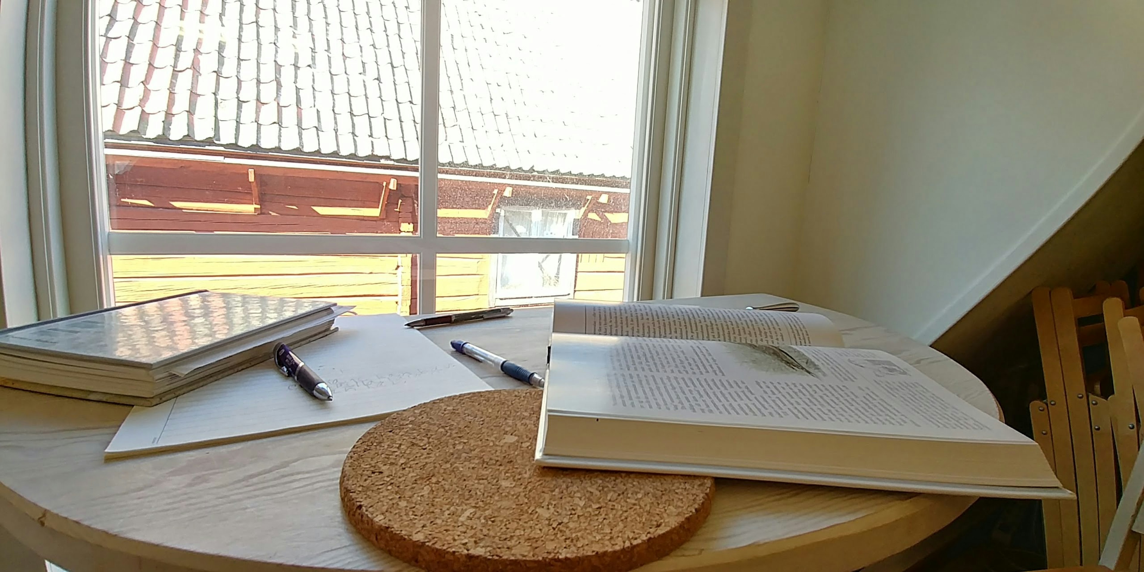 Wooden table near a window with books and pens and a cork coaster