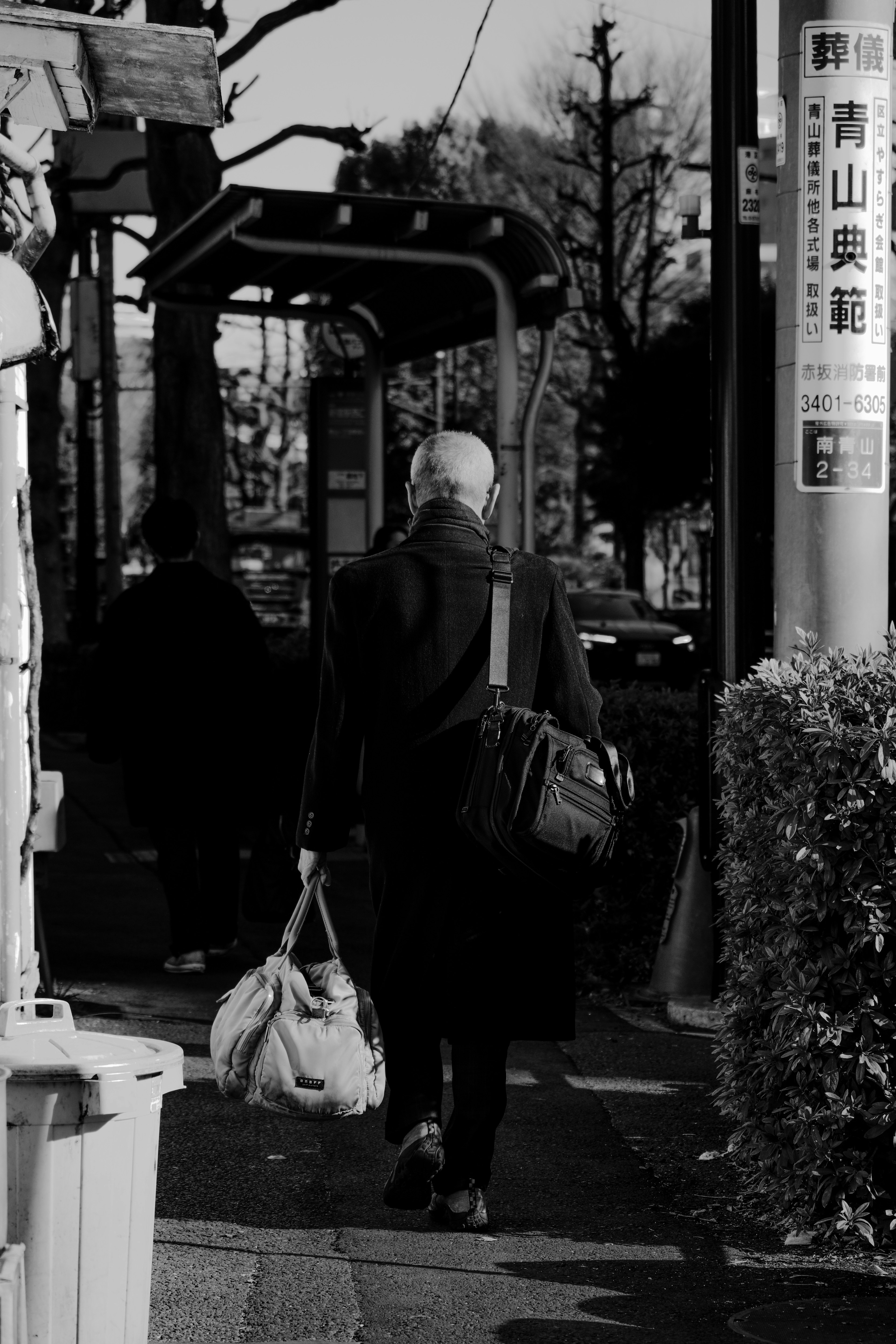 Foto in bianco e nero di un uomo con una borsa che cammina verso una fermata dell'autobus