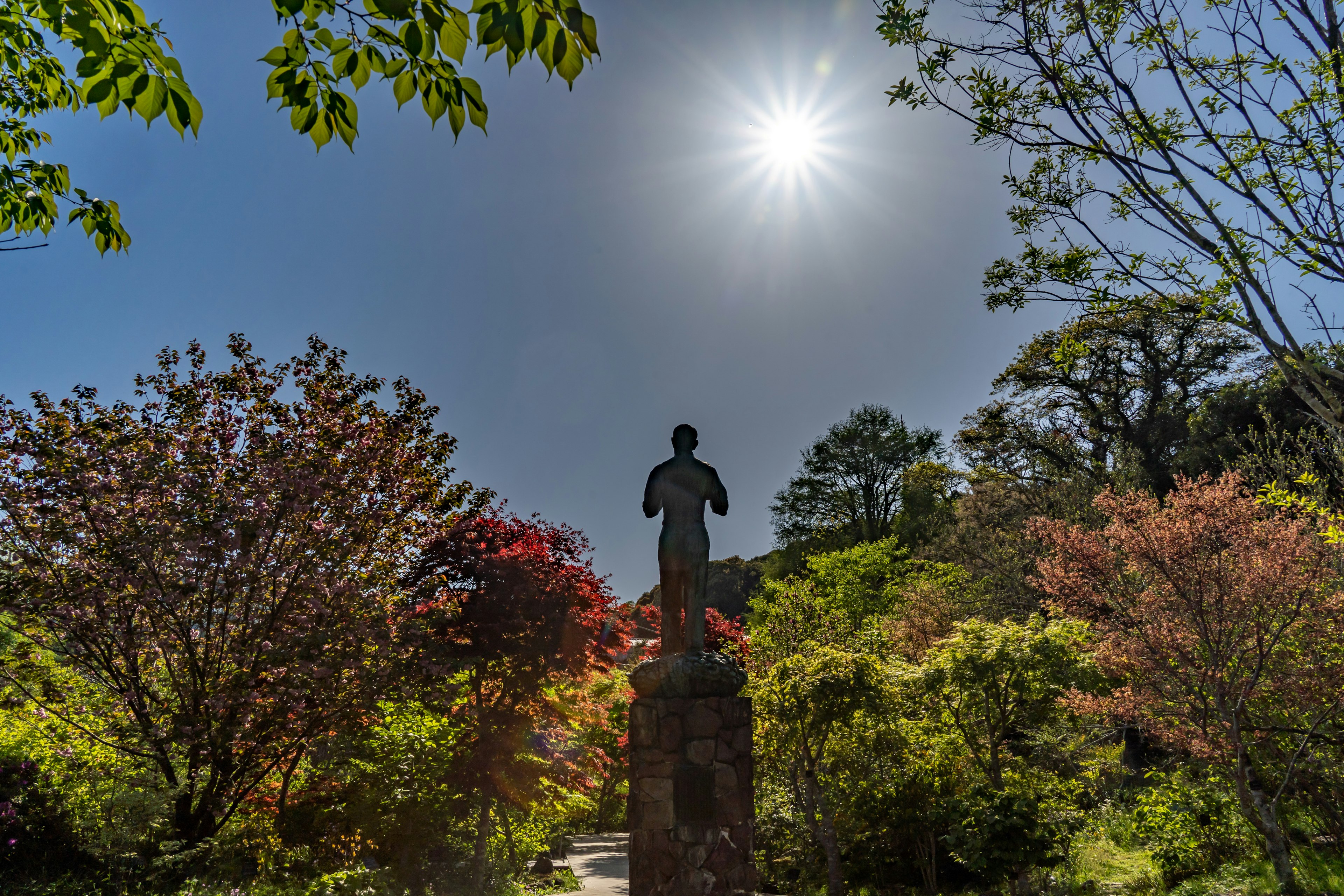 公園の中に立つ人物の像と色とりどりの木々を背景にした晴れた空