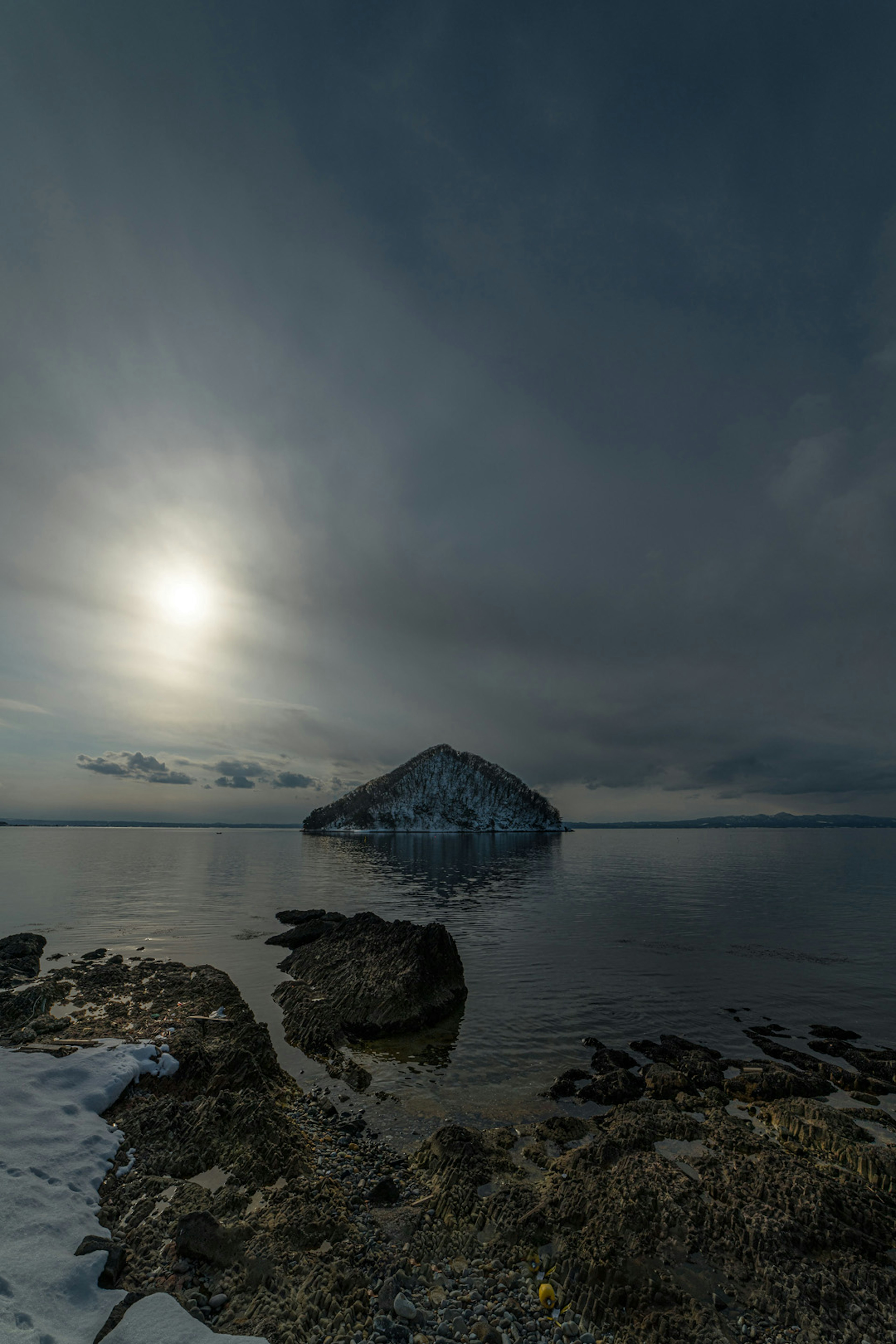 Un lago sereno con un'isola rocciosa innevata sotto un cielo nuvoloso