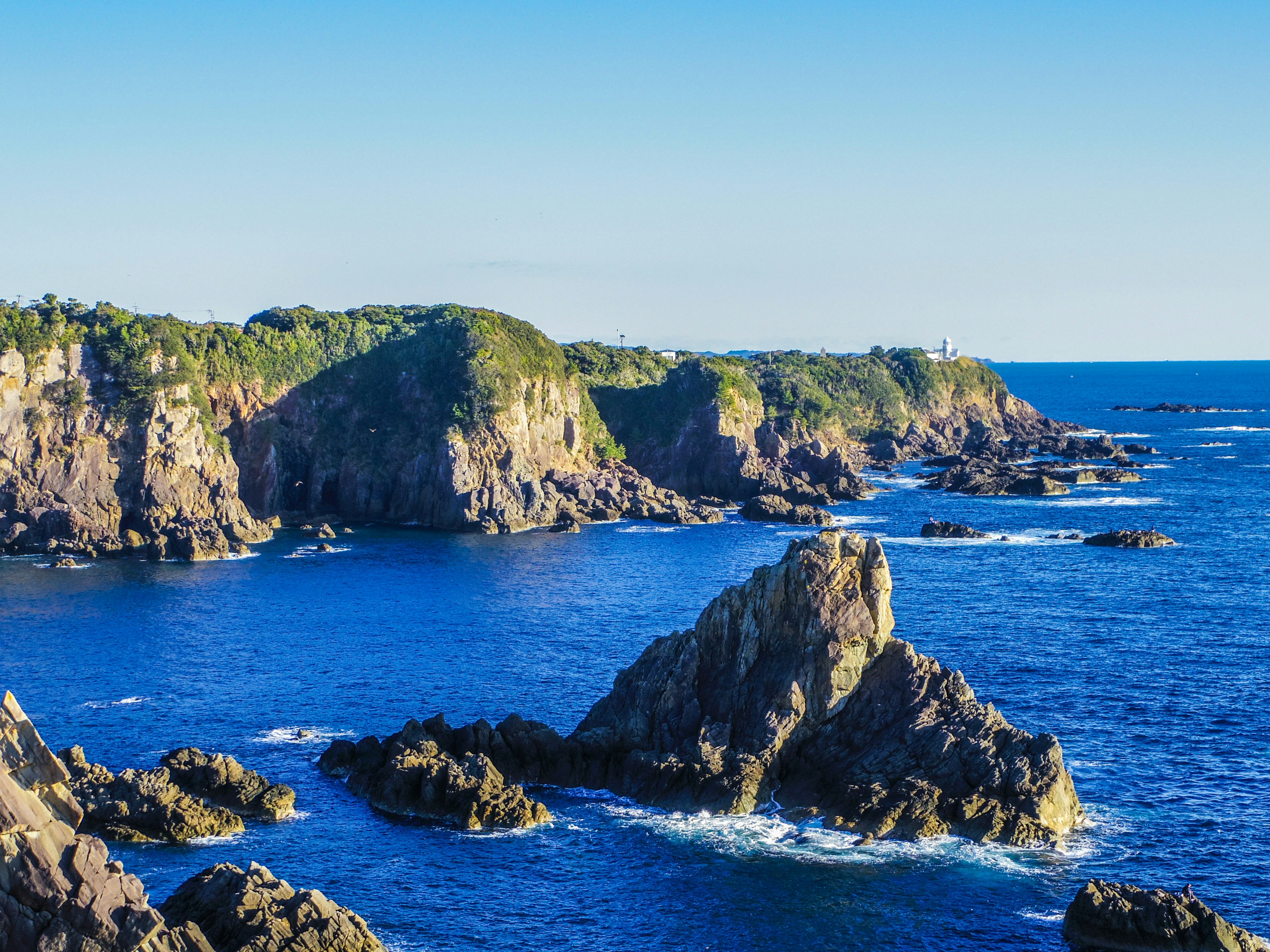 Vista costiera panoramica con oceano blu e scogliere rocciose con colline verdi e un faro in lontananza