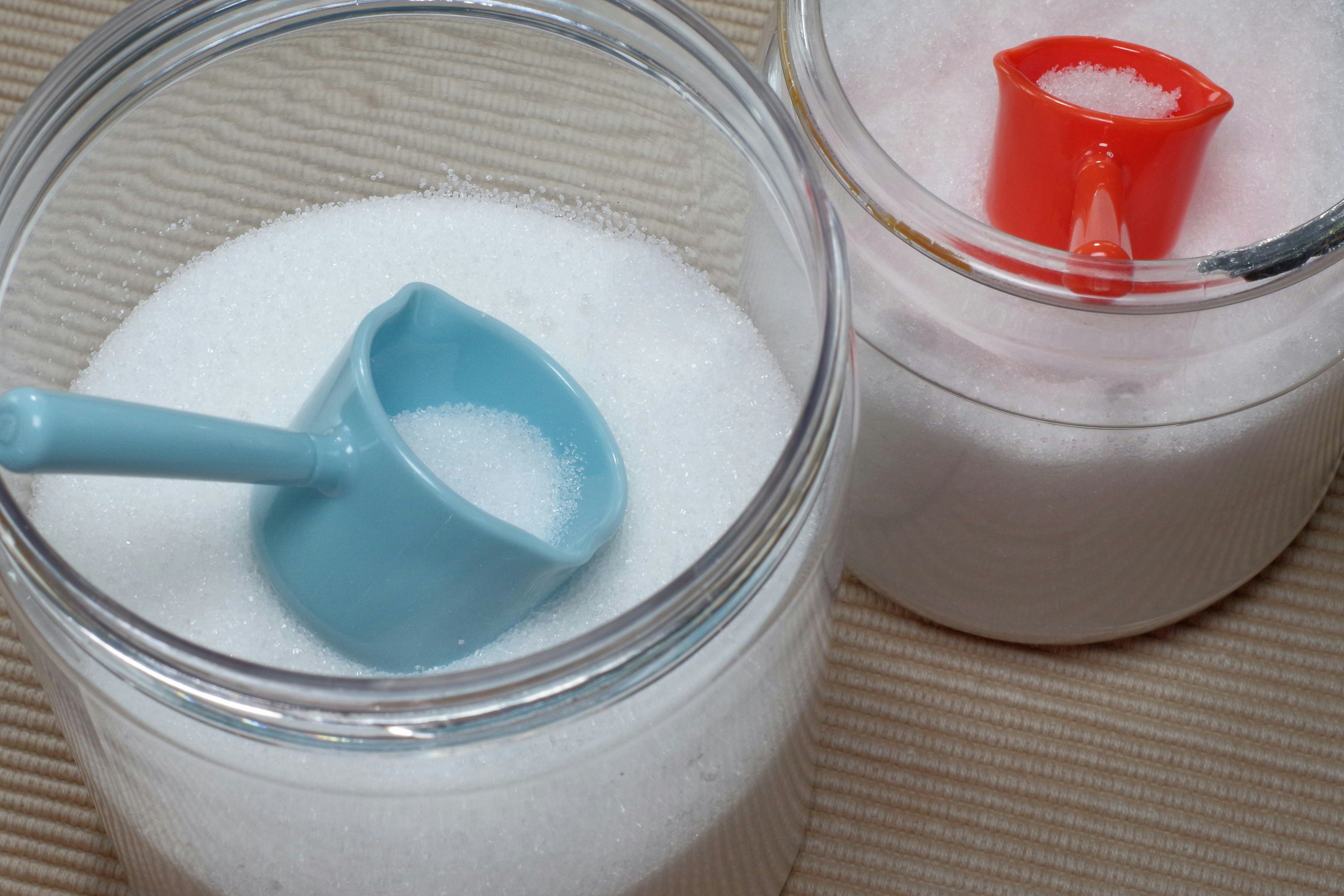 Two jars of sugar with a blue scoop and a red scoop