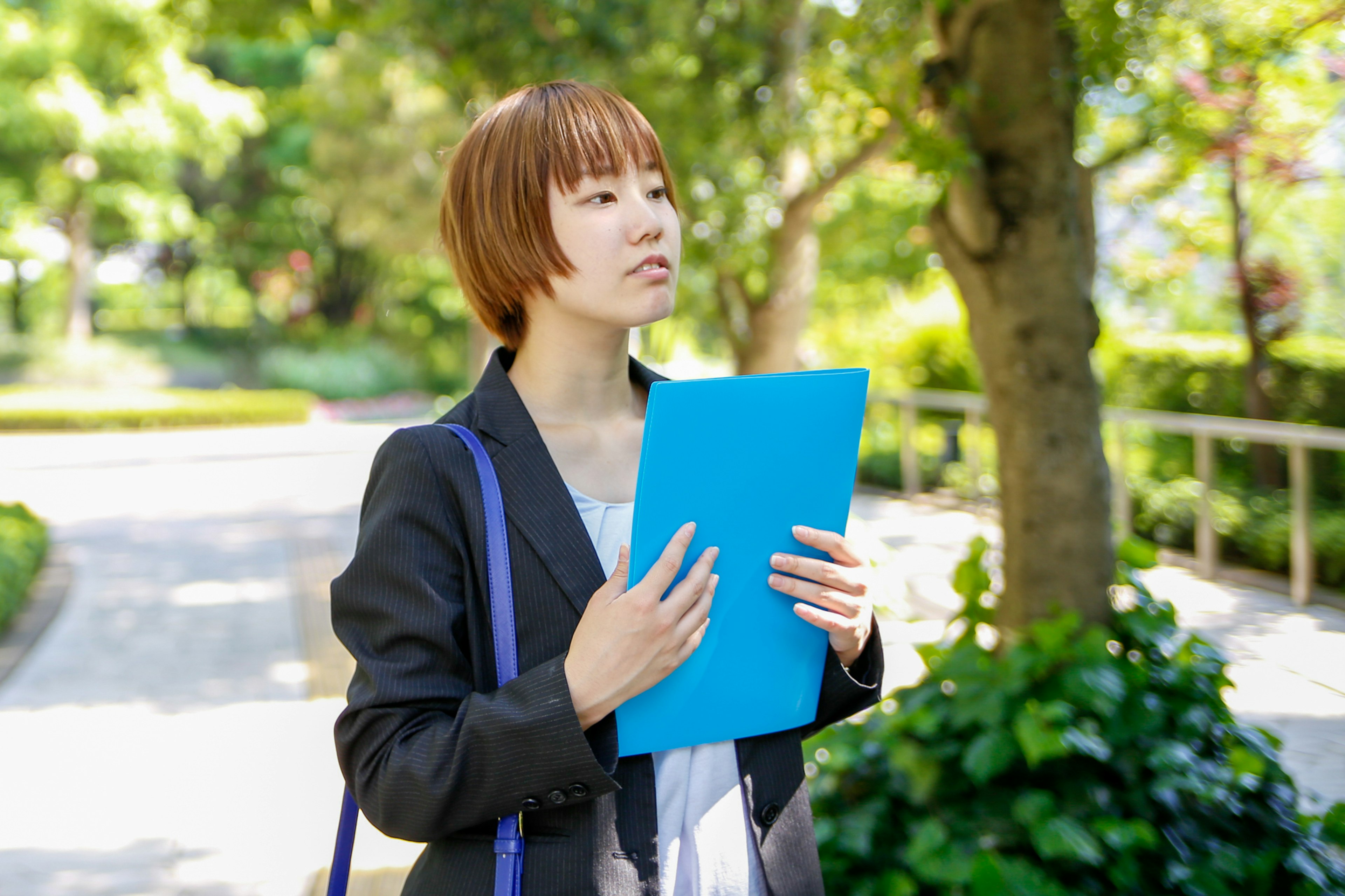 Young woman holding a blue folder looking thoughtful in a park