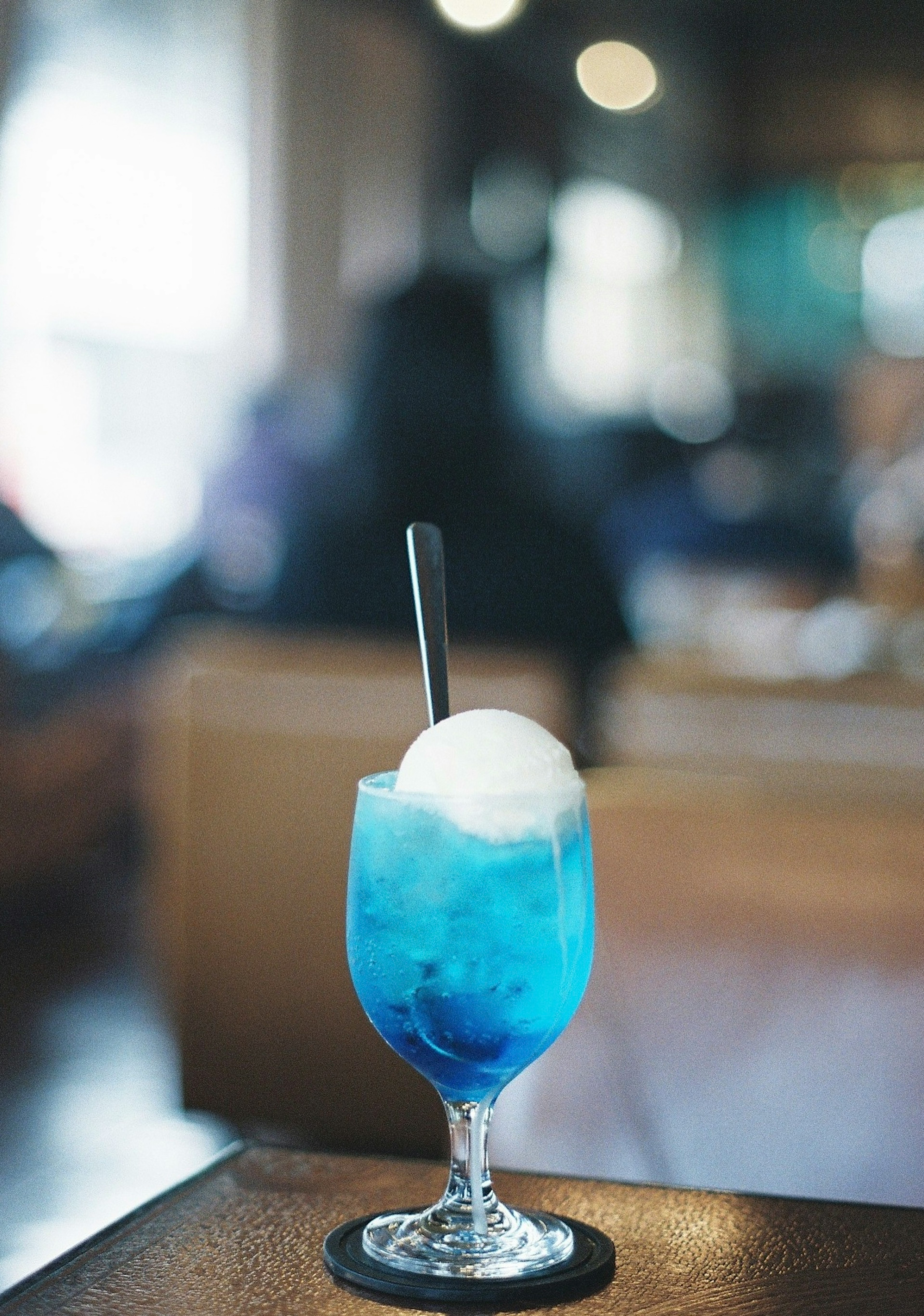 A blue drink topped with ice cream in a glass placed on a table with blurred people in the background