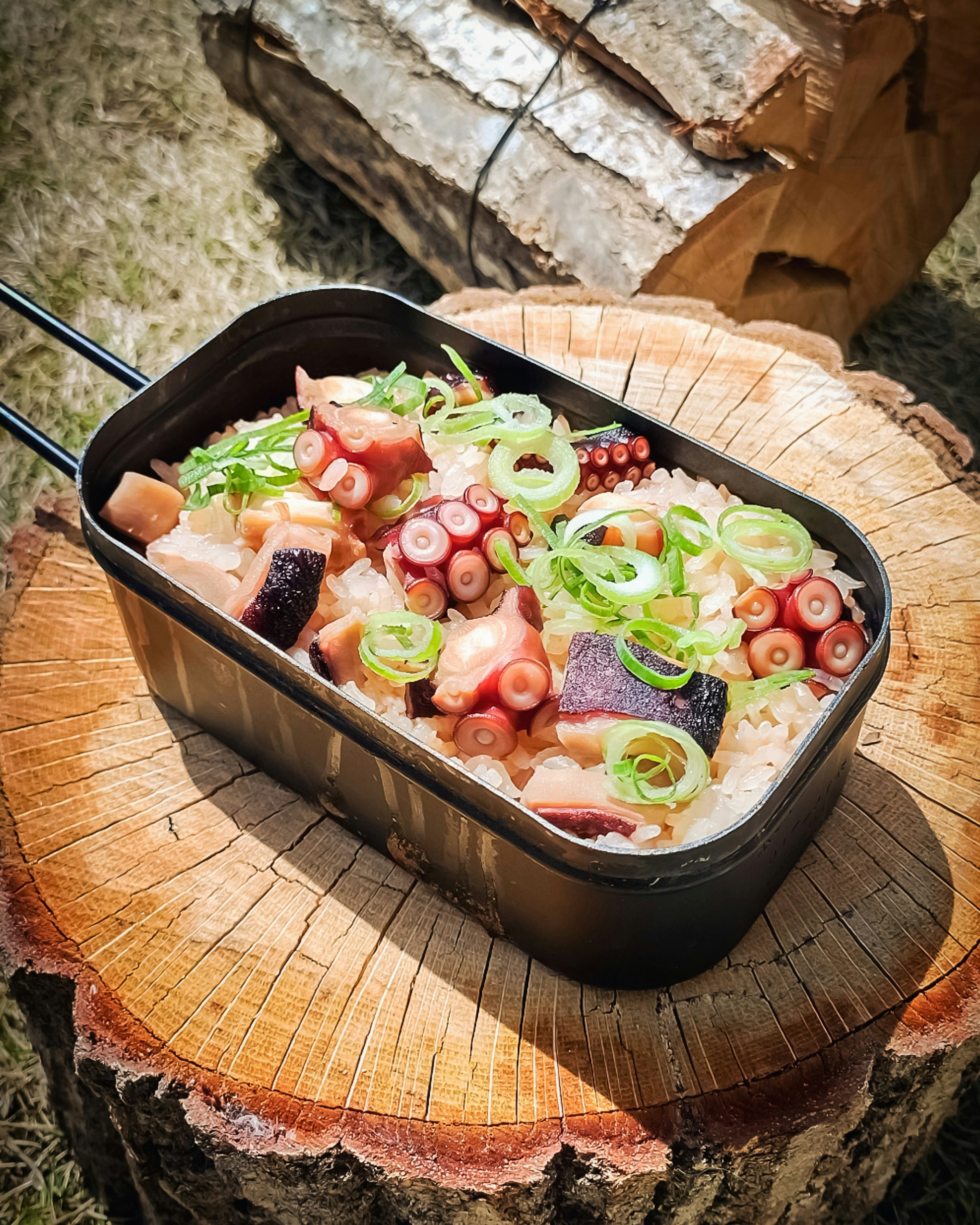 A black container filled with rice and octopus topped with green onions placed on a wooden stump