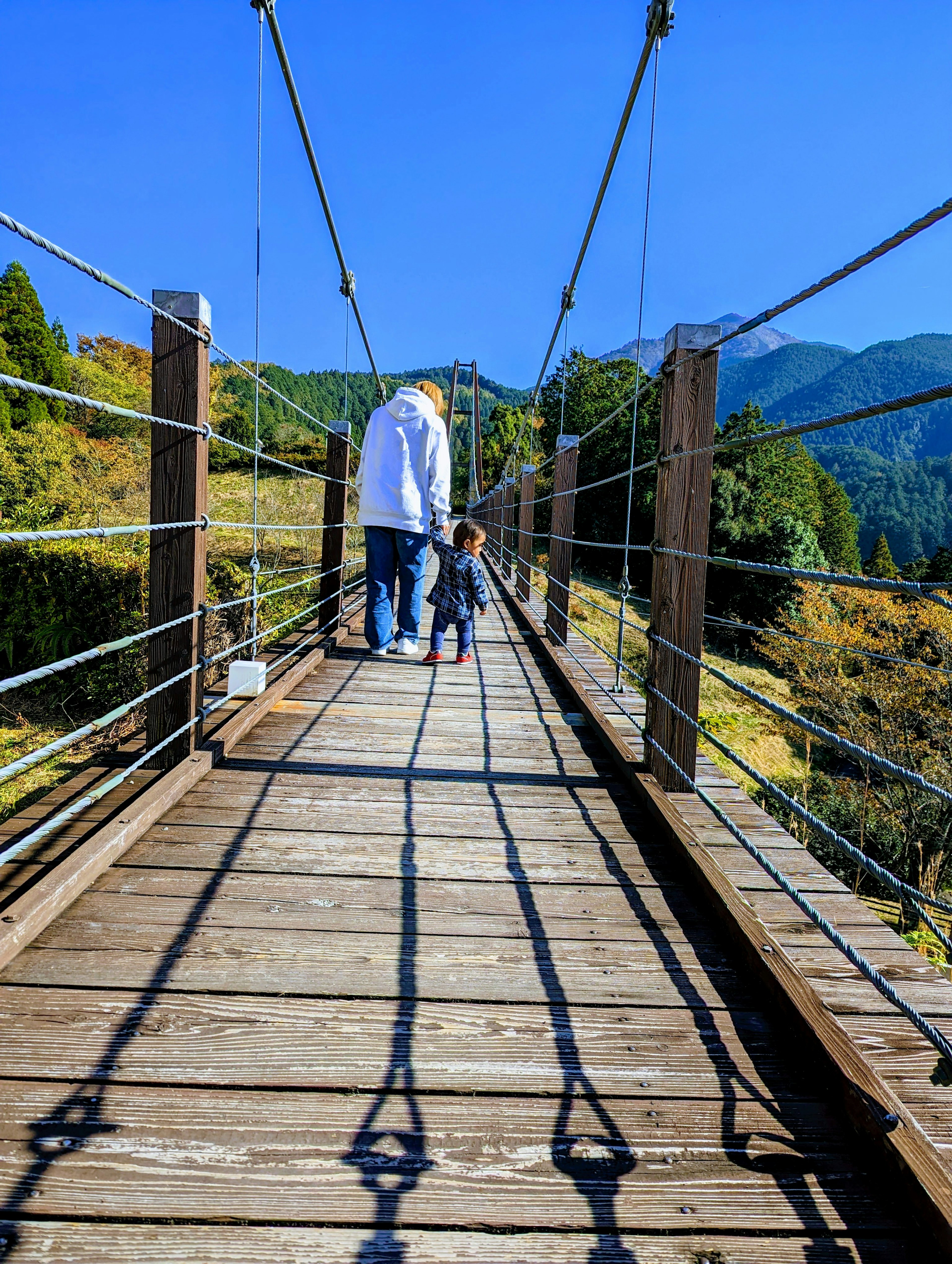 Adult and child walking on a suspension bridge with mountains and blue sky in the background striking shadows