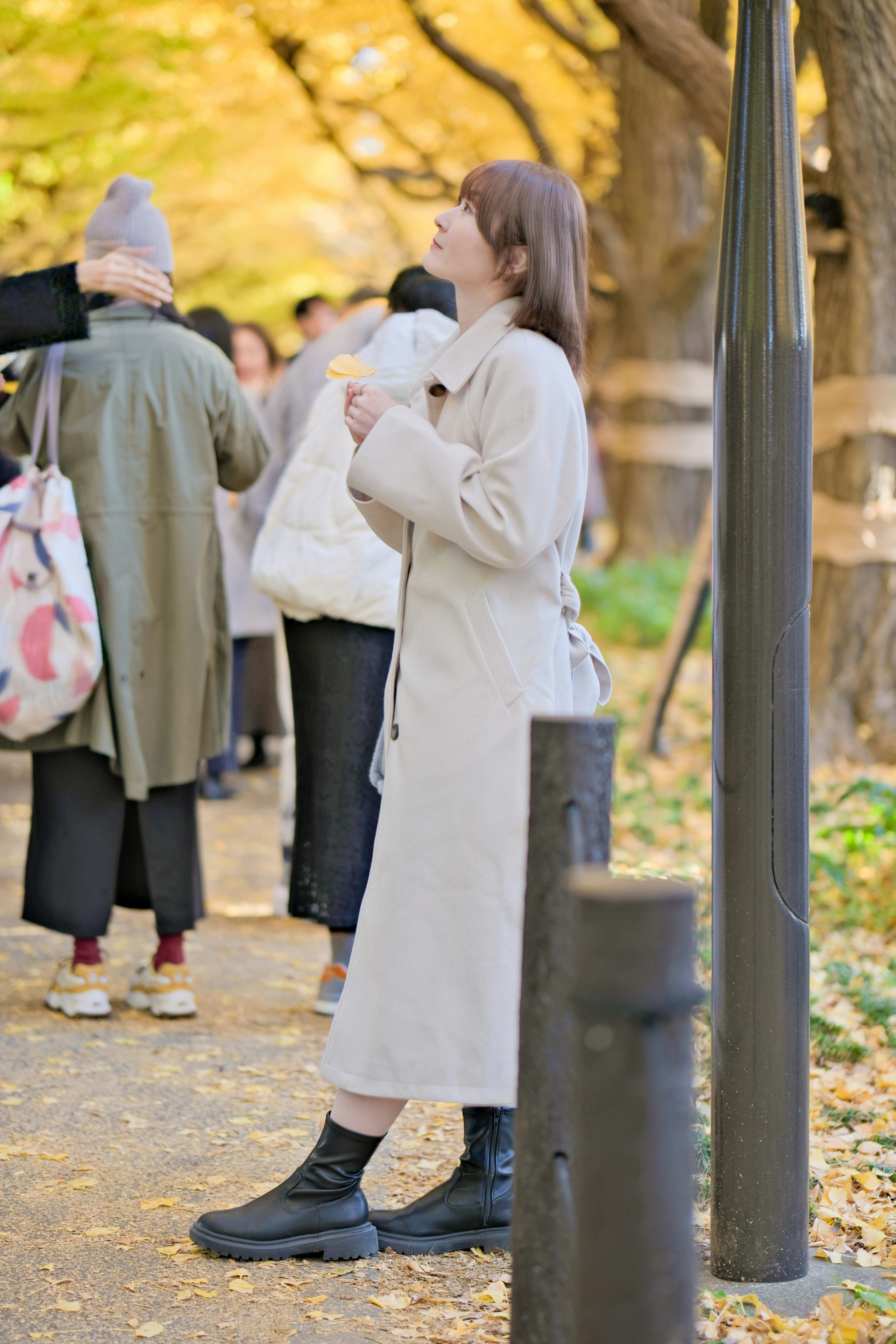 Una donna in piedi in un parco autunnale che indossa un cappotto bianco e stivali neri circondata da altre persone