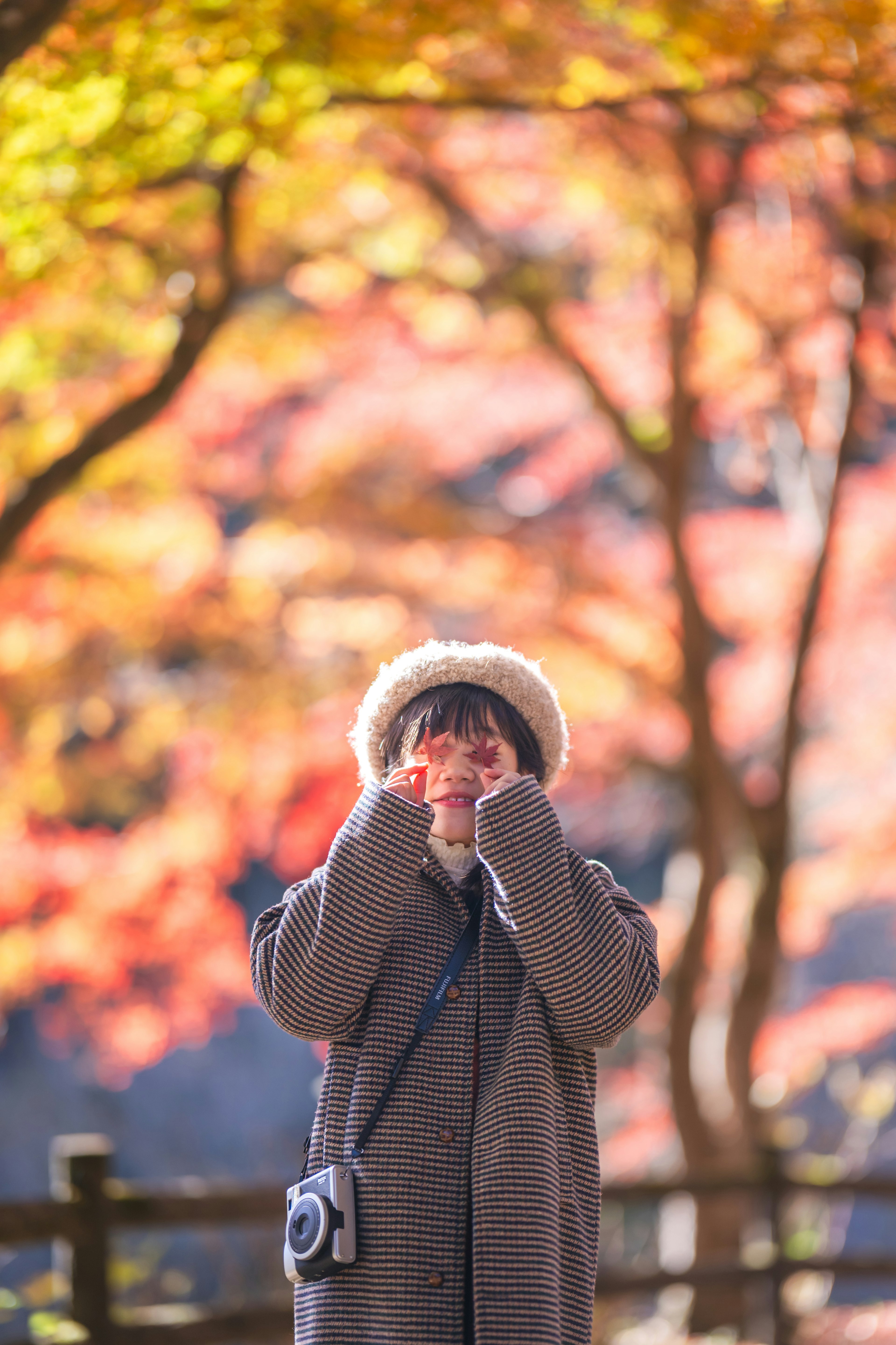 在秋天的树叶中摆姿势的女性穿着温暖的外套和帽子手持相机