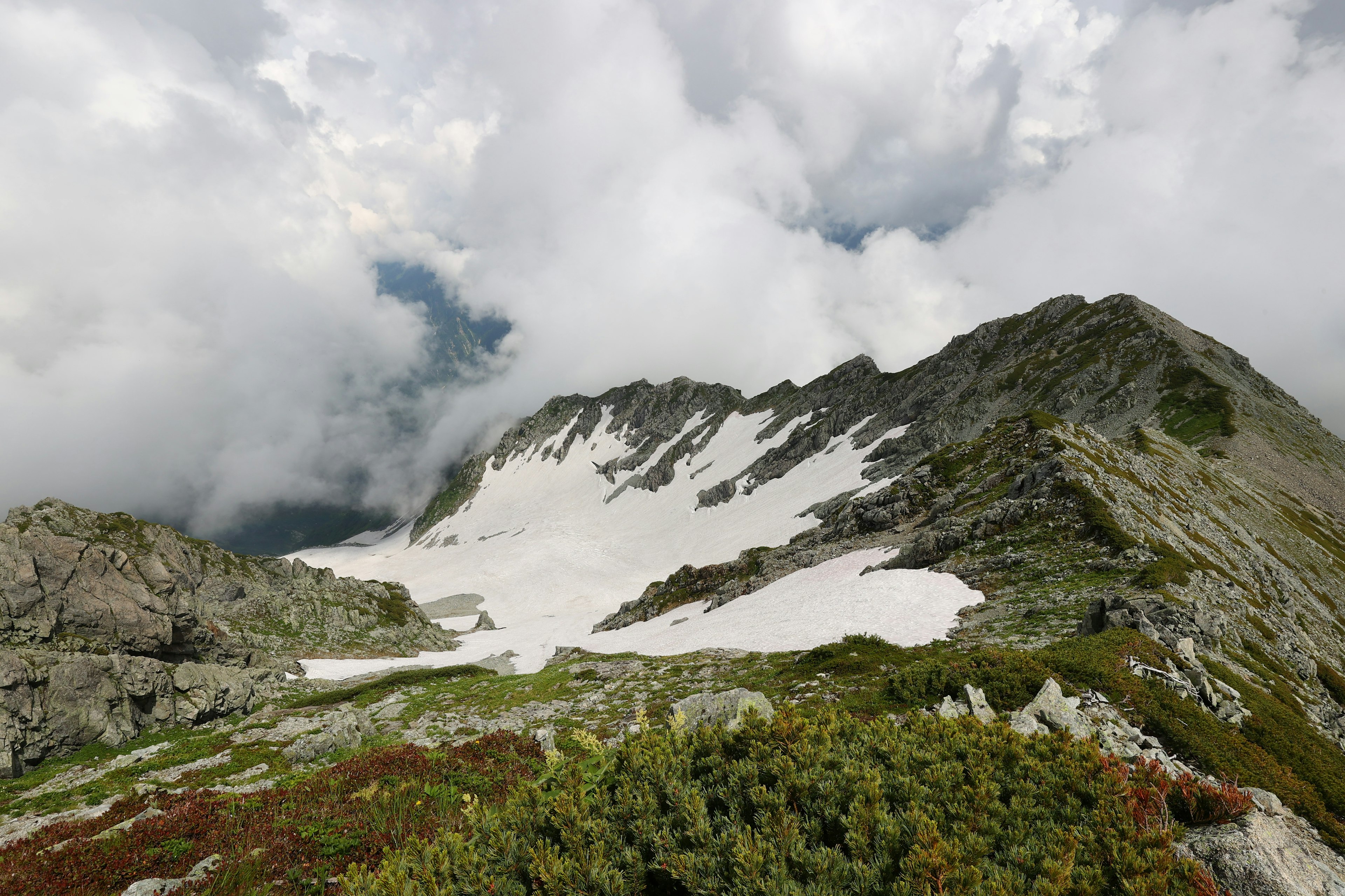 Vista de la cima de la montaña con nubes cubriendo las cumbres y parches de nieve rodeados de vegetación verde