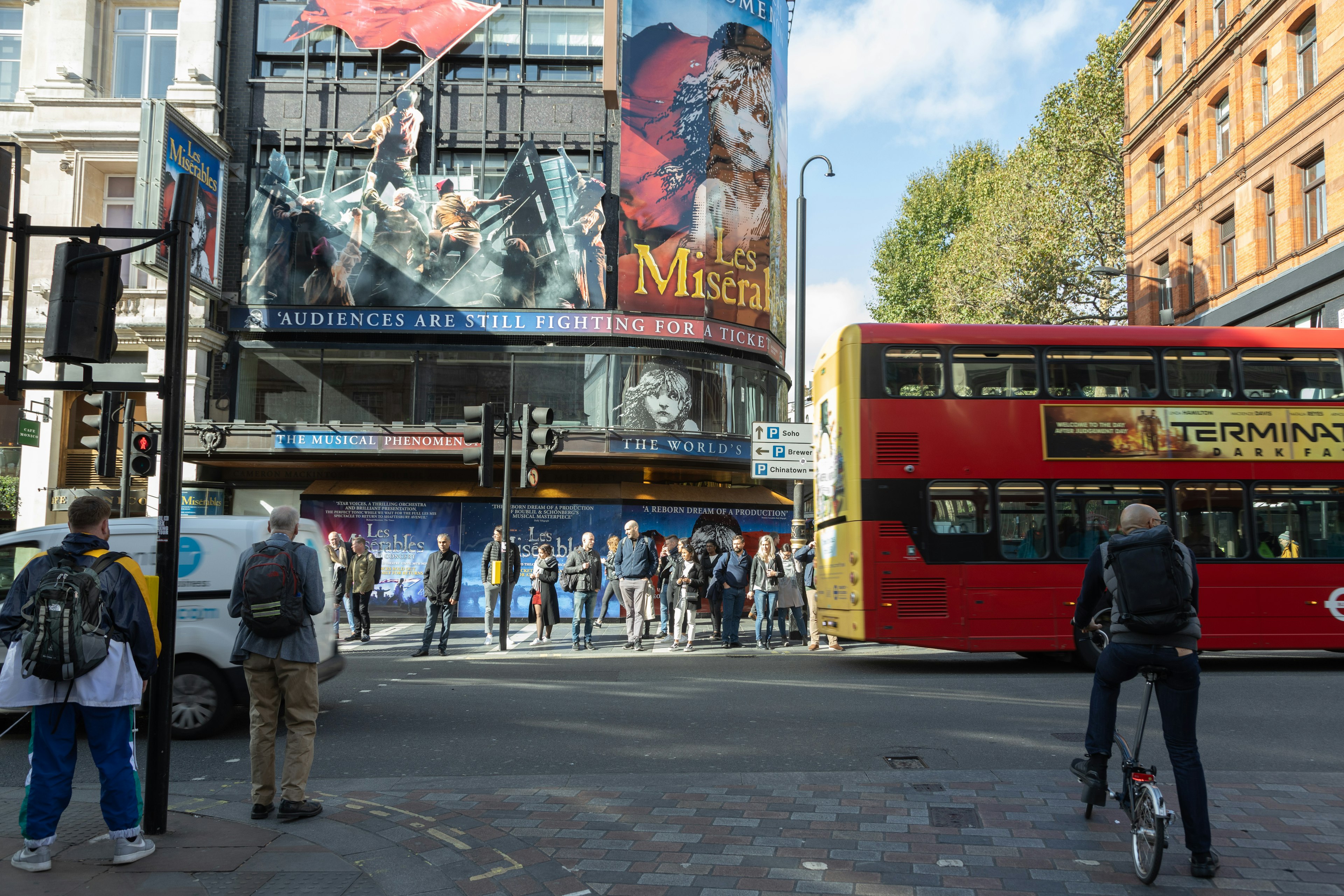 Szene einer belebten Londoner Straße mit einem Musical-Werbeschild an einem Gebäude und einem roten Bus