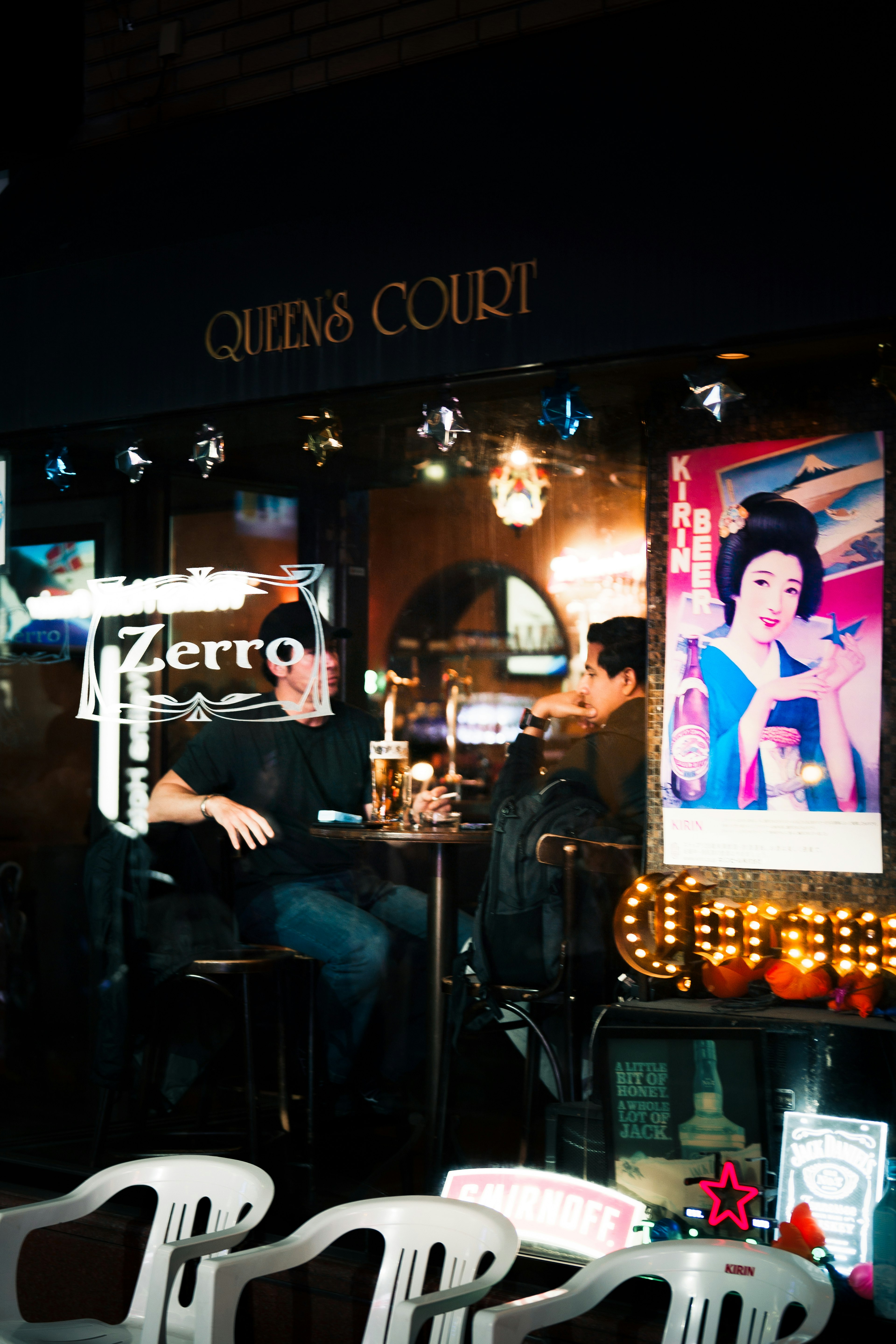 Zwei Männer sitzen in einer Bar mit bunten Schildern im Hintergrund