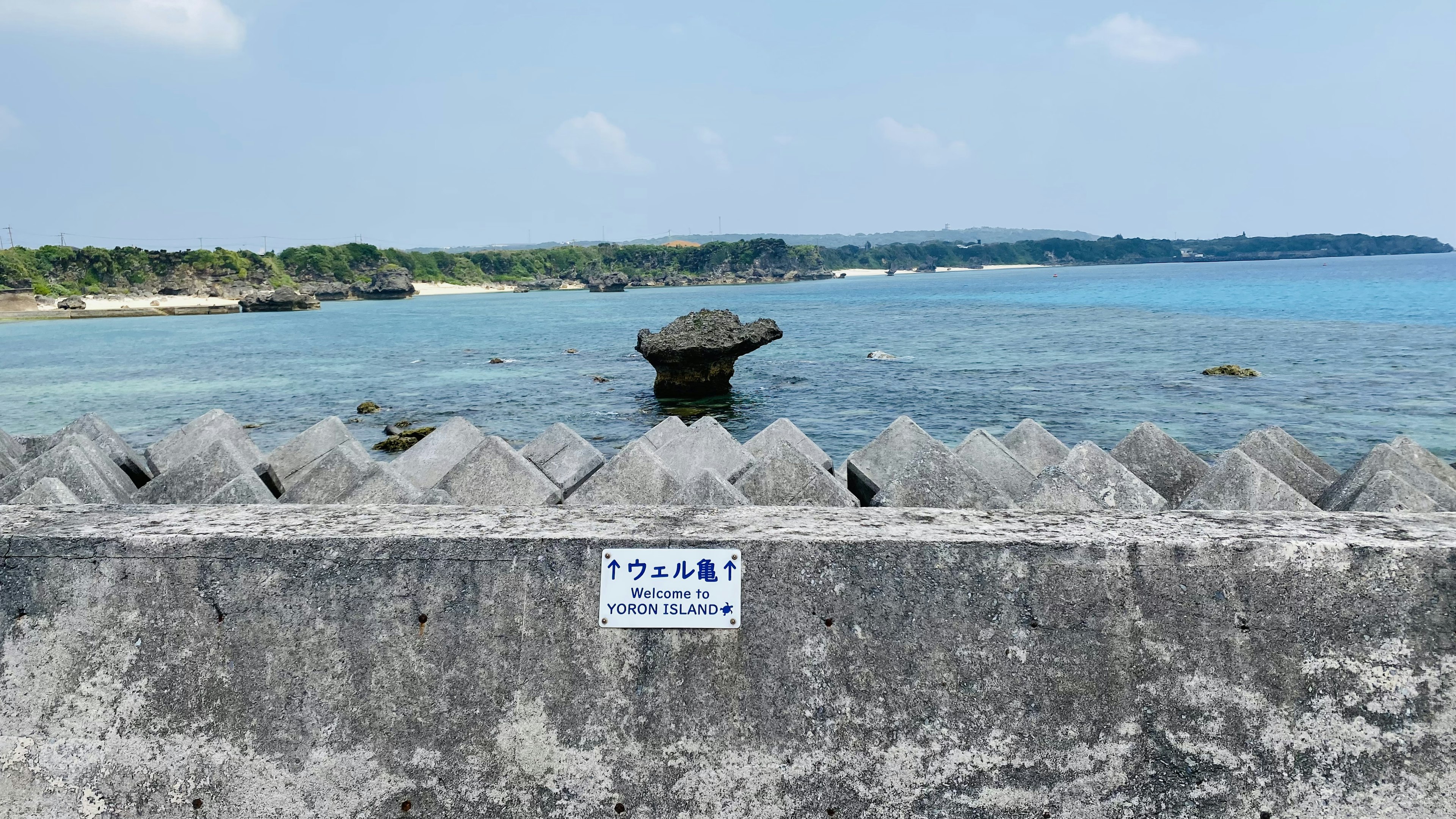 Landscape image featuring the sea and a breakwater with a stone object in the center and a blue sky and coastline in the background