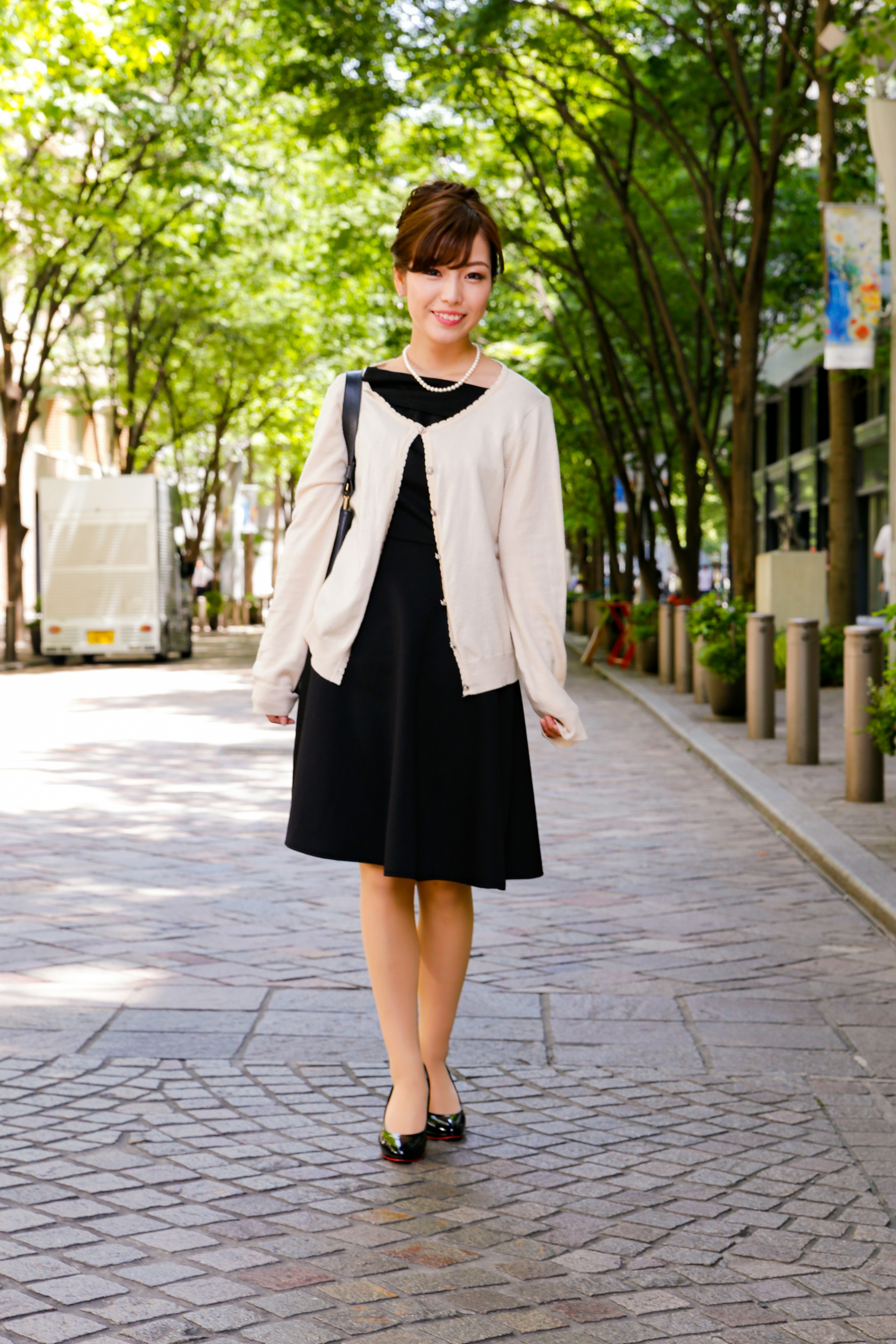 Una donna in un vestito nero e un cardigan bianco che cammina in una strada alberata