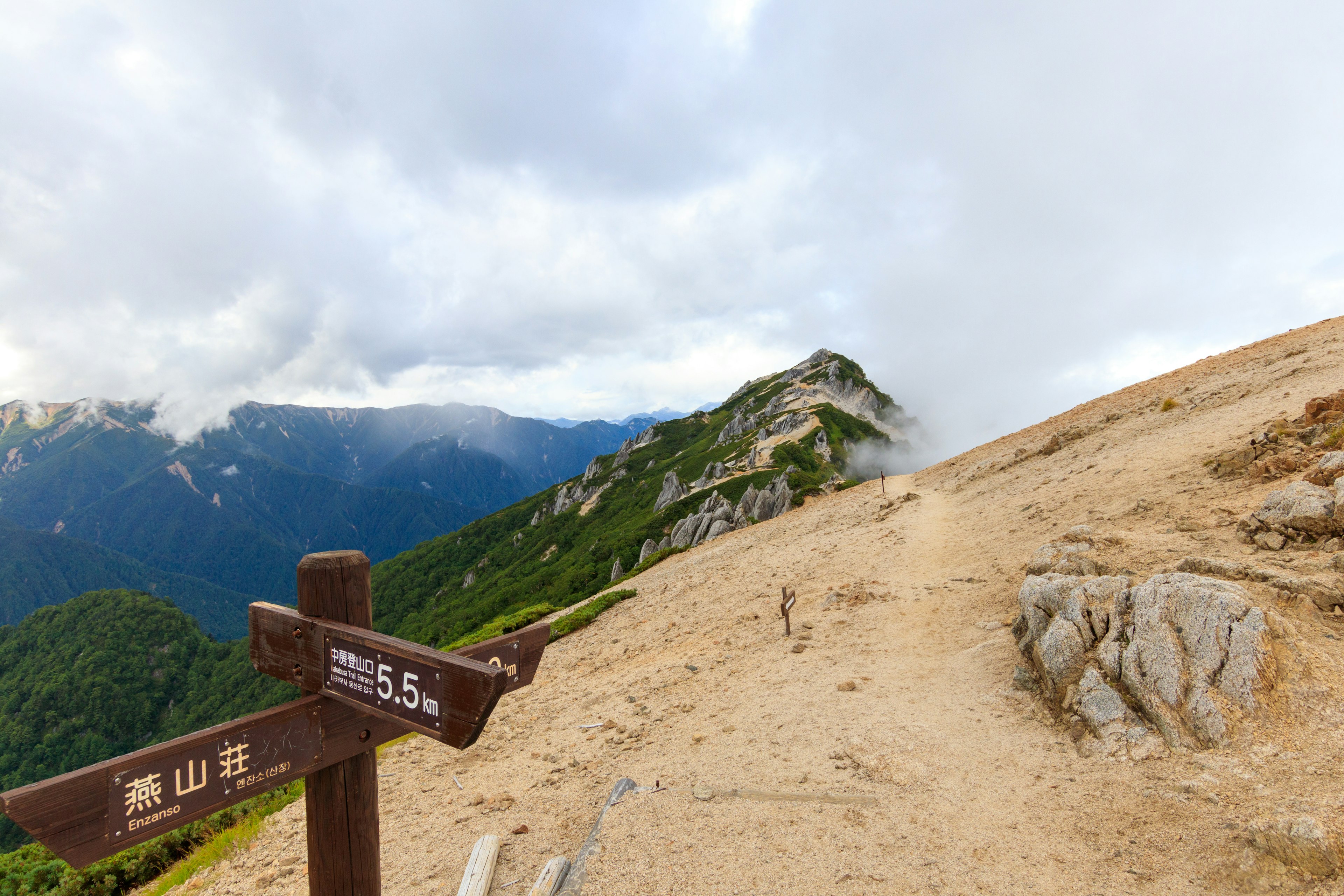 Sendero montañoso con un letrero que indica 5,5 kilómetros hasta la cima rodeado de nubes y terreno accidentado